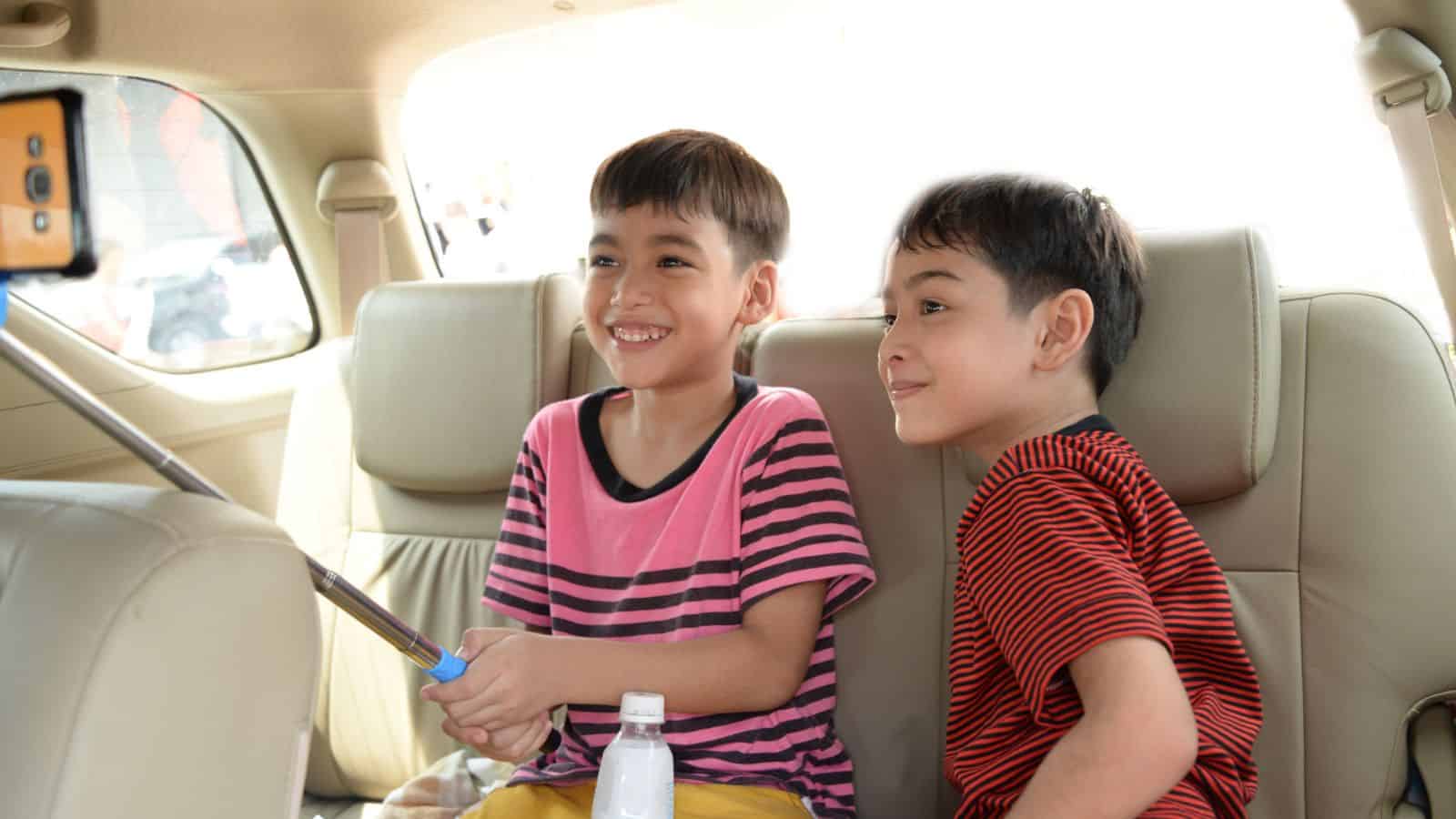 Two young boys sit in a car’s backseat, smiling and taking a selfie with a smartphone on a selfie stick.
