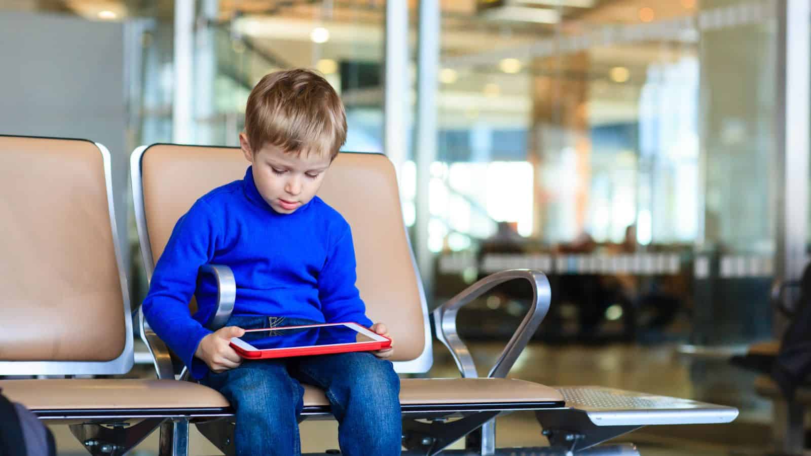 Young boy in a blue shirt sitting at an airport, looking at a tablet on his lap.