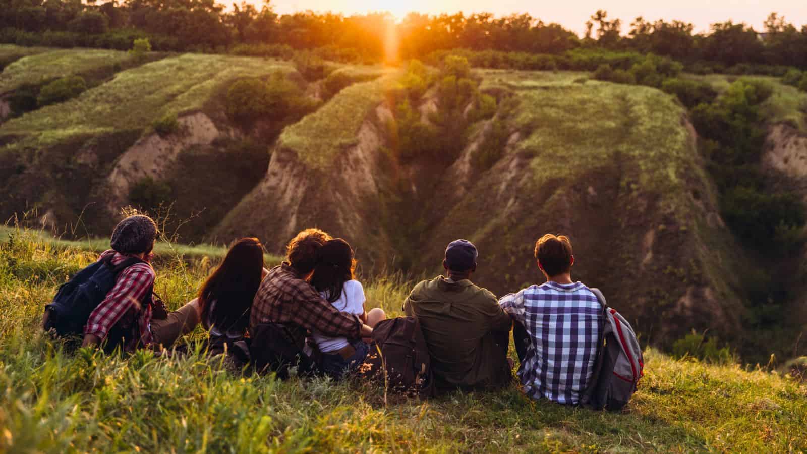 Six people sit on a grassy hill, watching the sunset over a green valley with trees and cliffs.