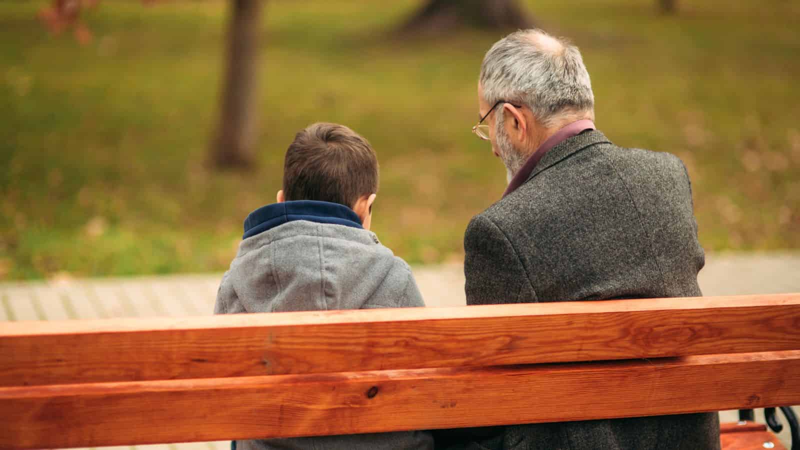 An elderly man and a young boy sit together on a wooden bench in a park, seen from behind.
