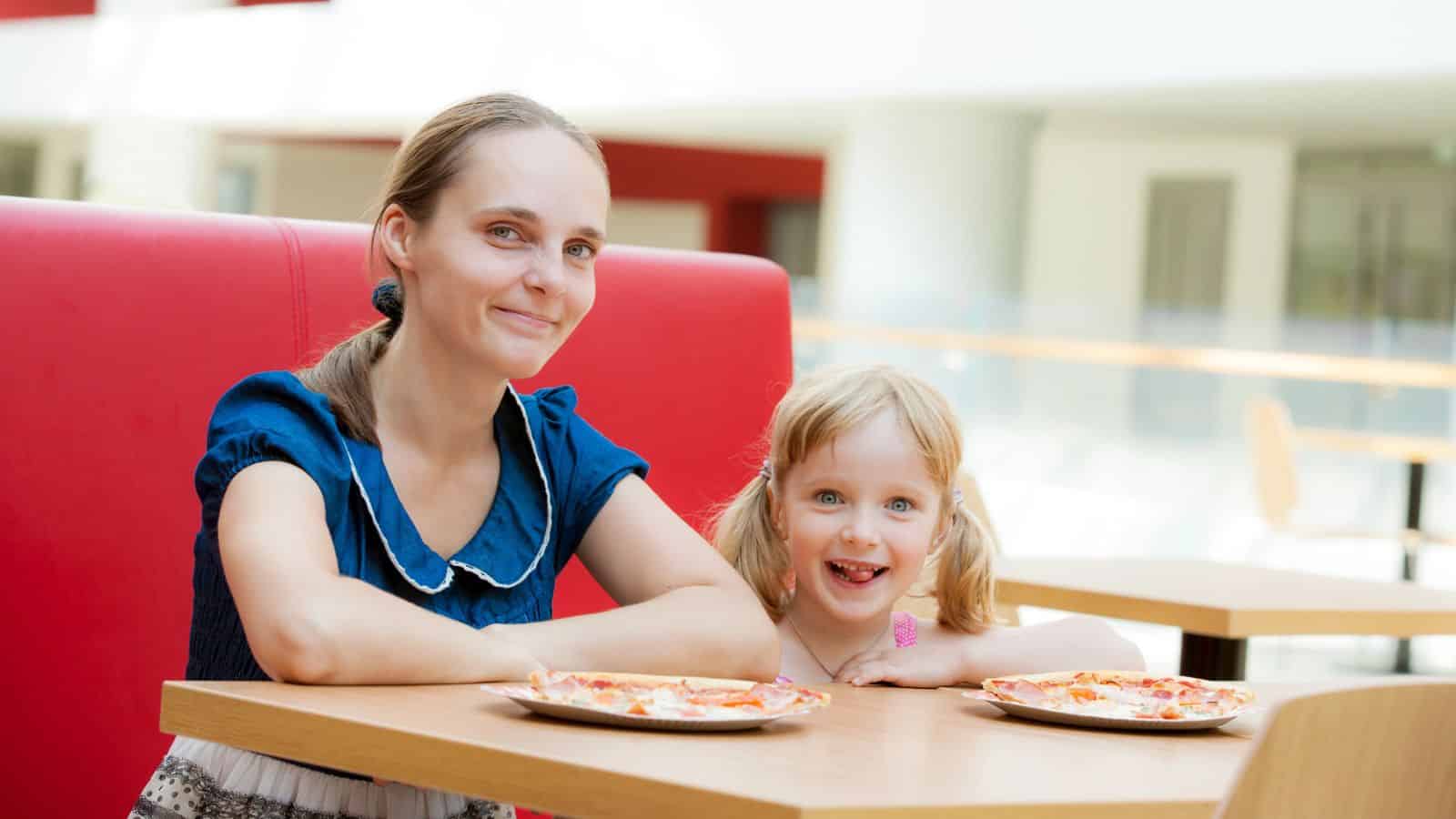 A woman and a young girl smile at a table with pizza in a bright, modern restaurant.