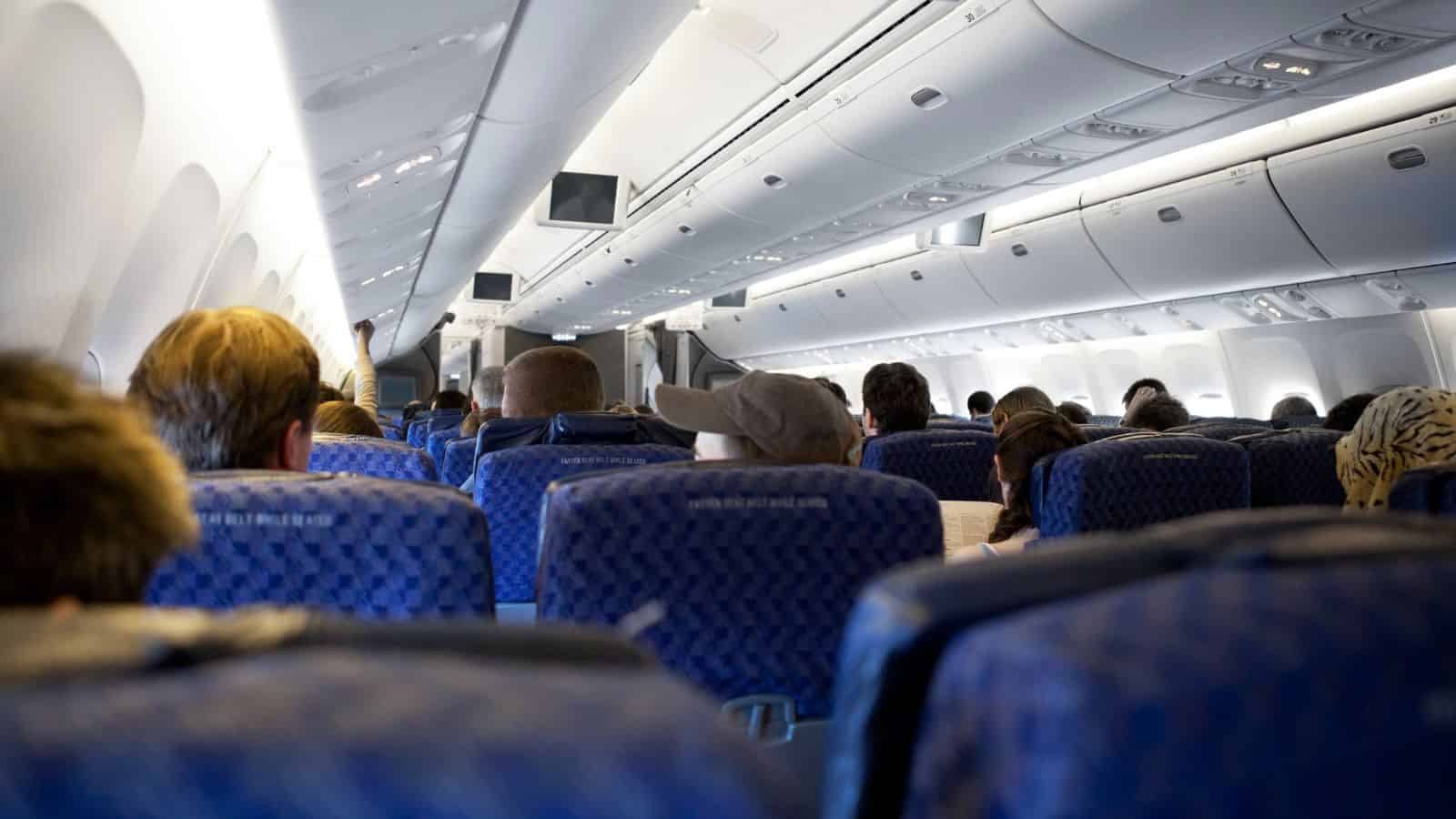 Passengers seated inside a commercial airplane cabin, viewed from the back toward the front aisle.