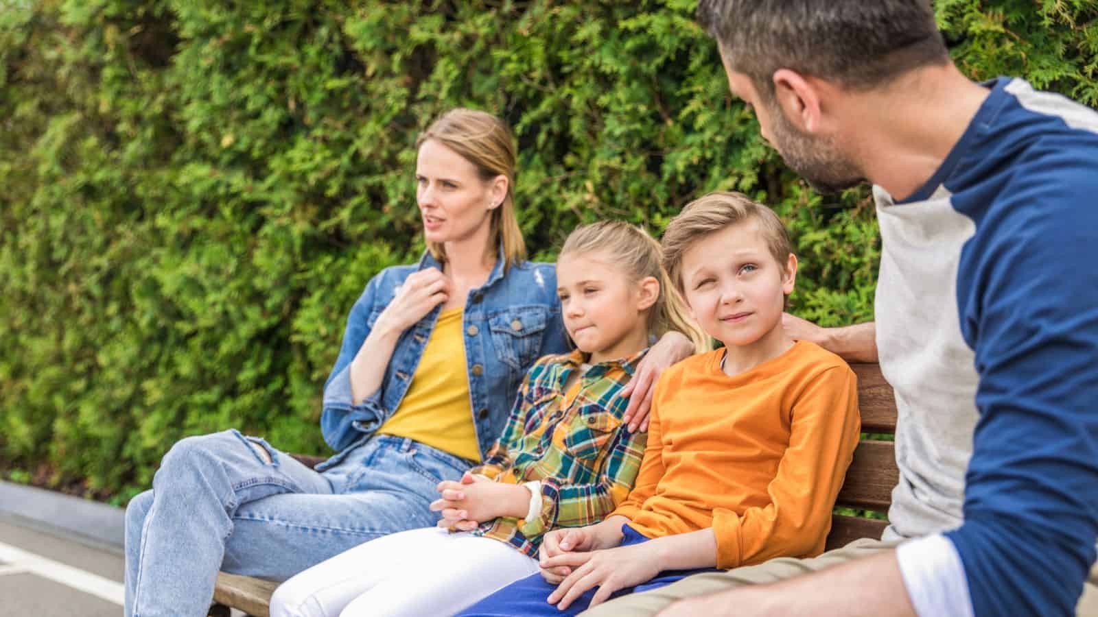 Four people, two adults and two children, sit on a bench outdoors, talking and enjoying the day.