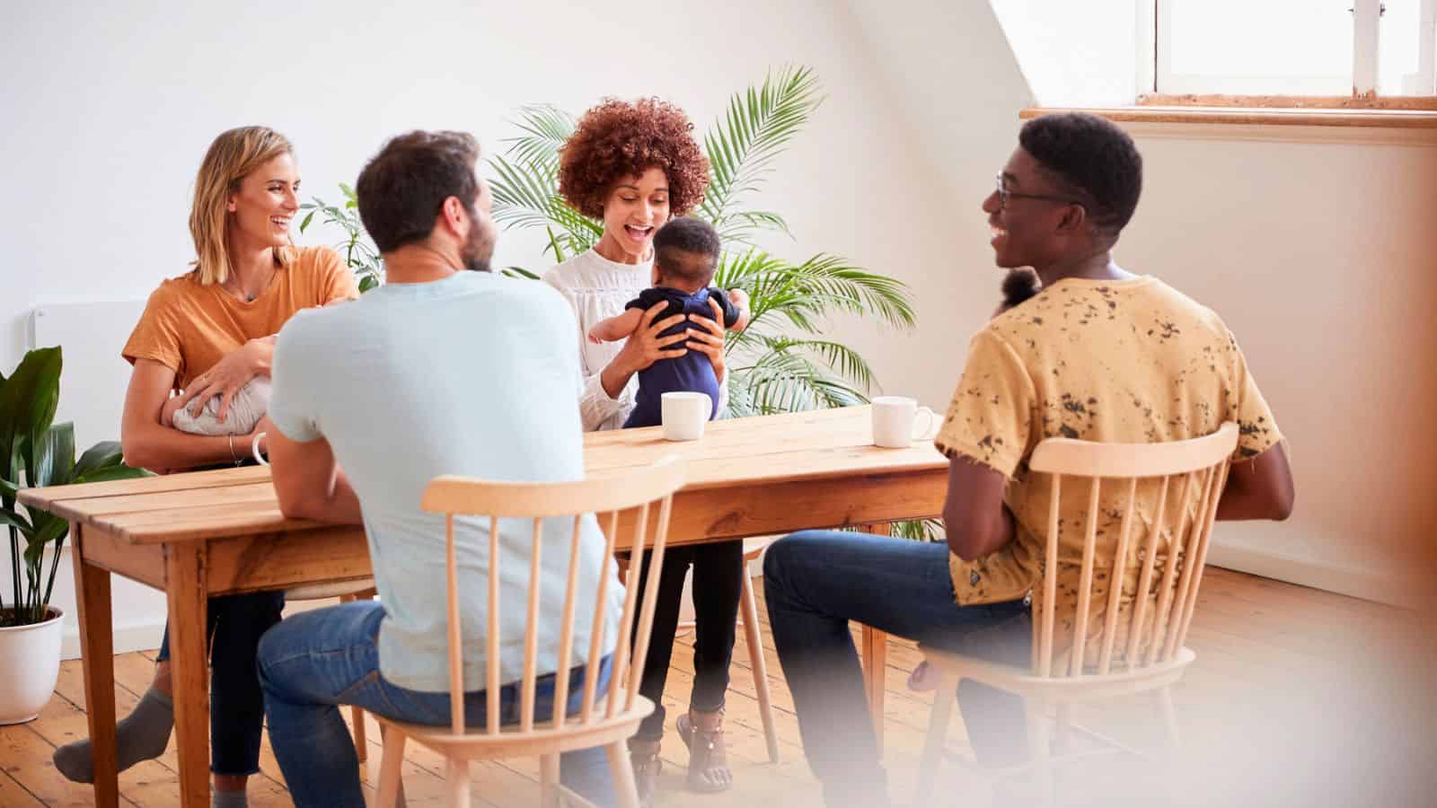 Four adults sit around a wooden table, smiling, as one woman holds a baby in a cozy, sunlit room.