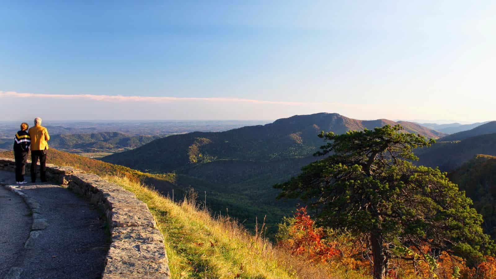 Two people stand by a stone wall overlooking scenic mountains under a clear blue sky.