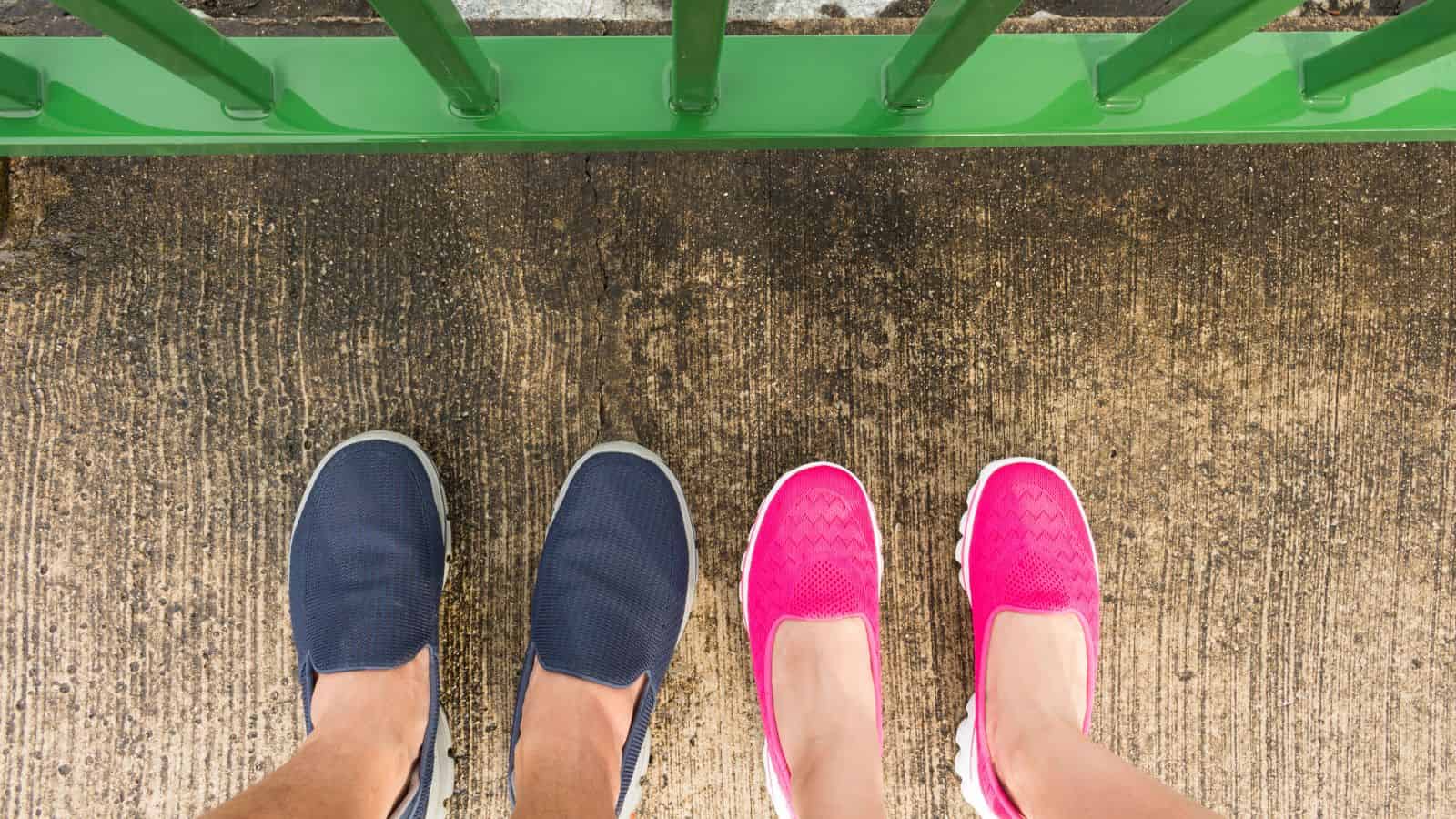 Two people standing on pavement, one wearing blue shoes and the other wearing bright pink shoes, near a green railing.