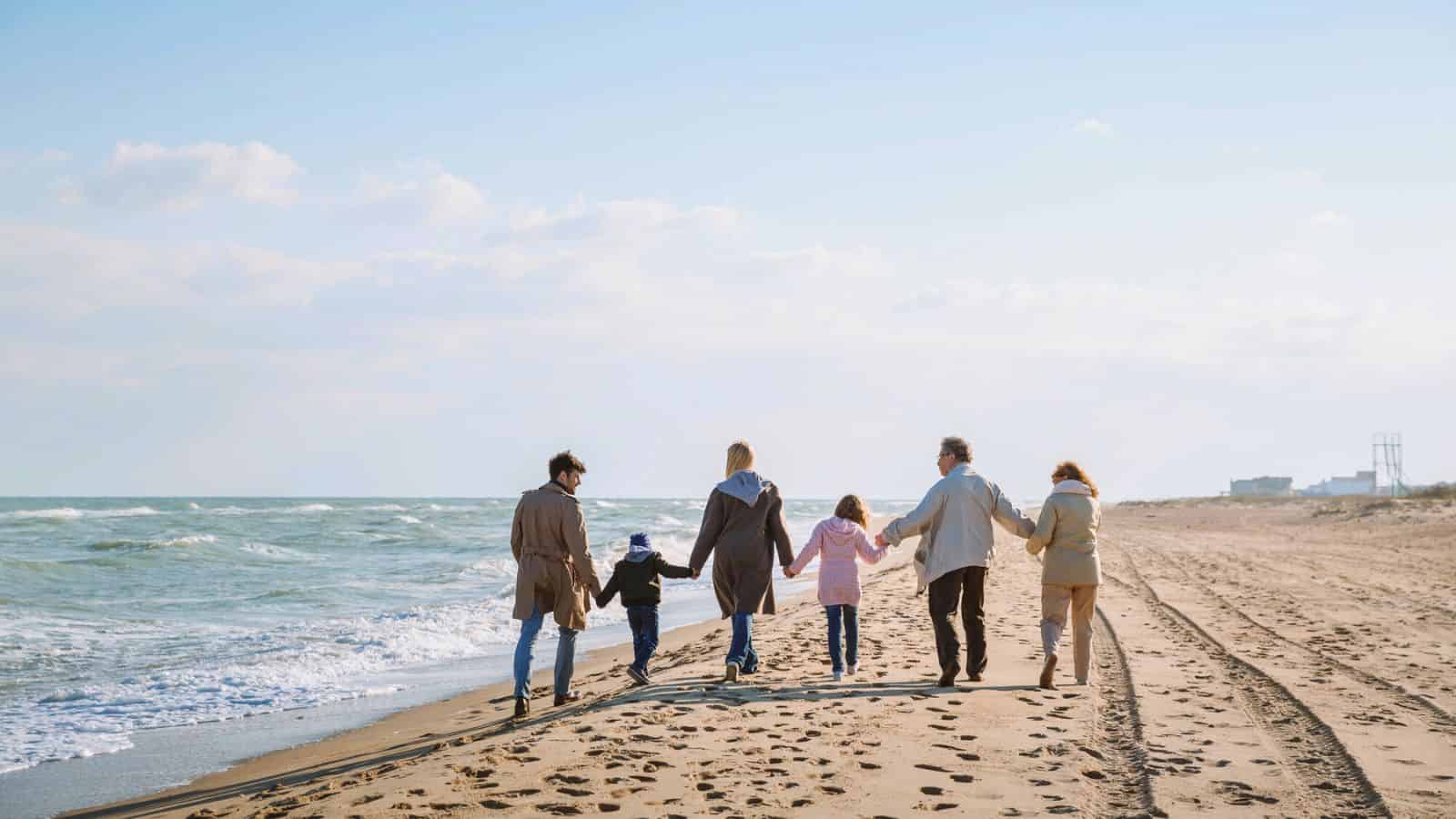 Six people holding hands walking along a sandy beach by the ocean on a clear, sunny day.