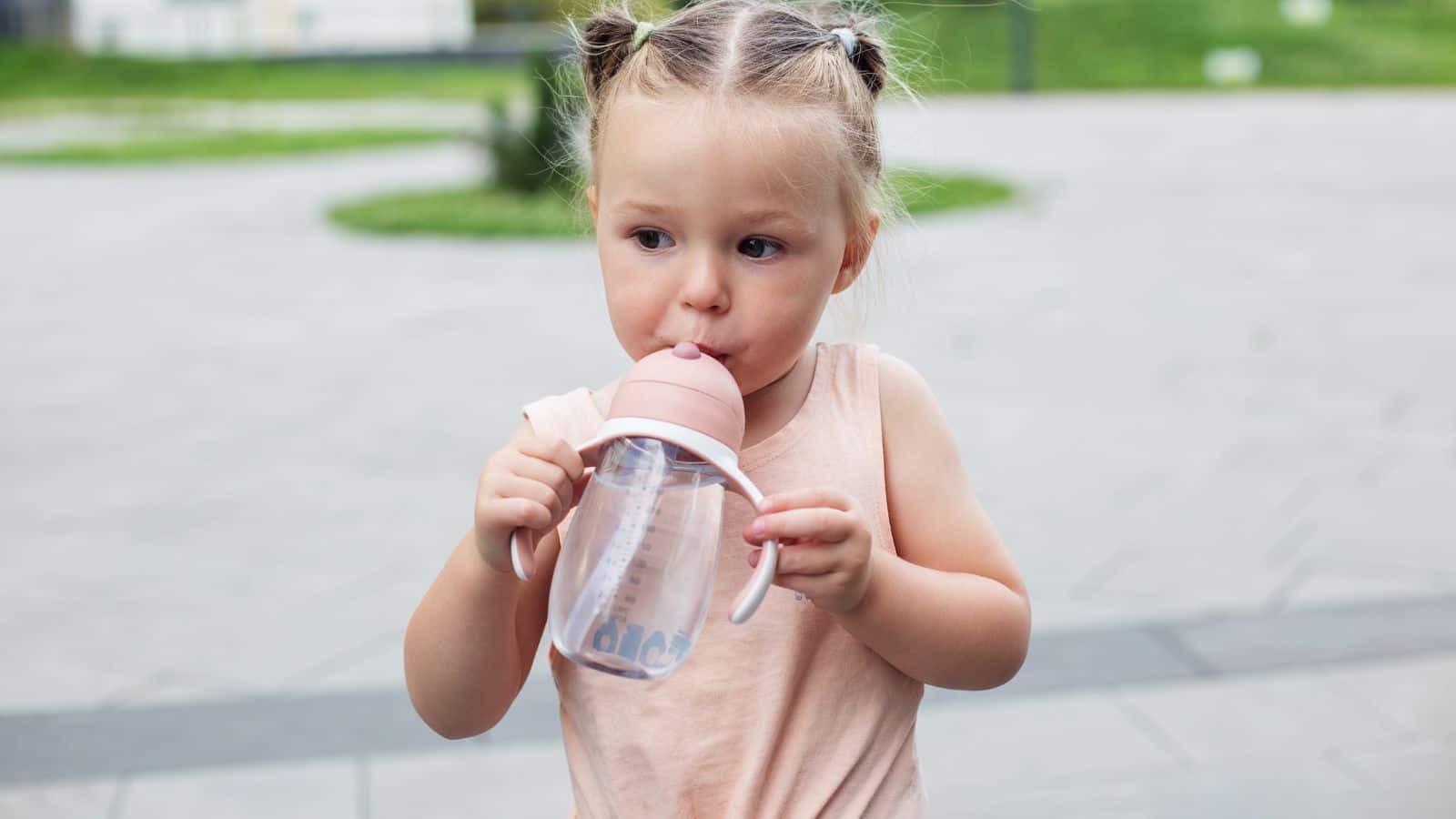 Young girl with pigtails drinking from a sippy cup while standing outdoors on a paved area—a perfect snapshot of travel essentials for kids under 5.