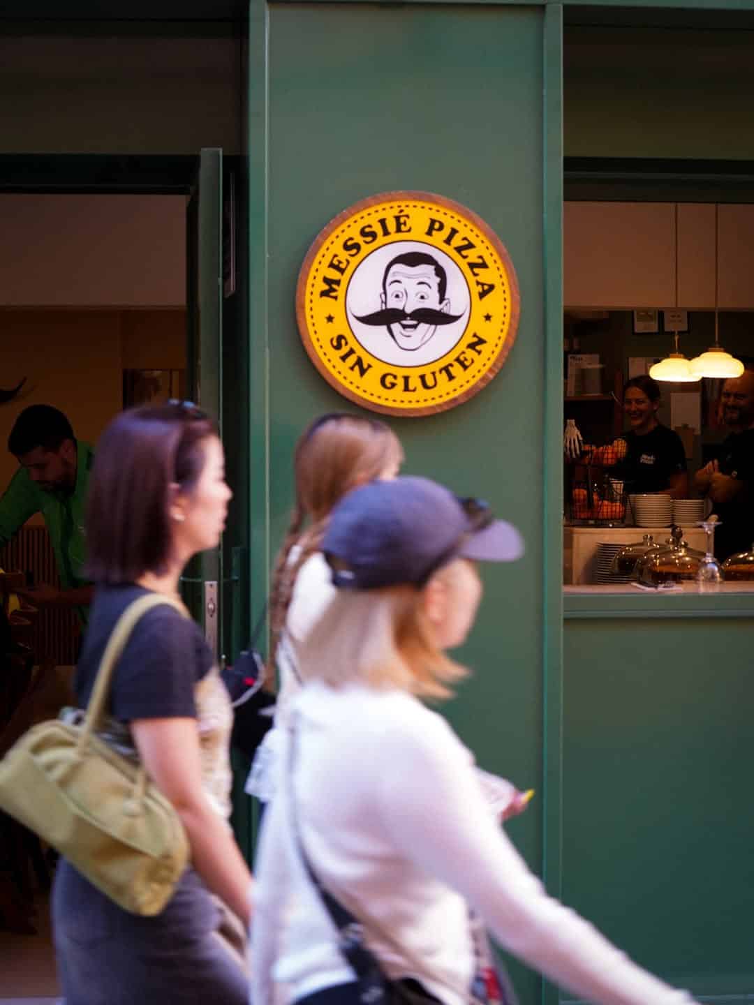 People walk past a green pizza shop in Gluten Free Barcelona, marked by a round "Messie Pizza Sin Gluten" sign featuring a smiling chef.