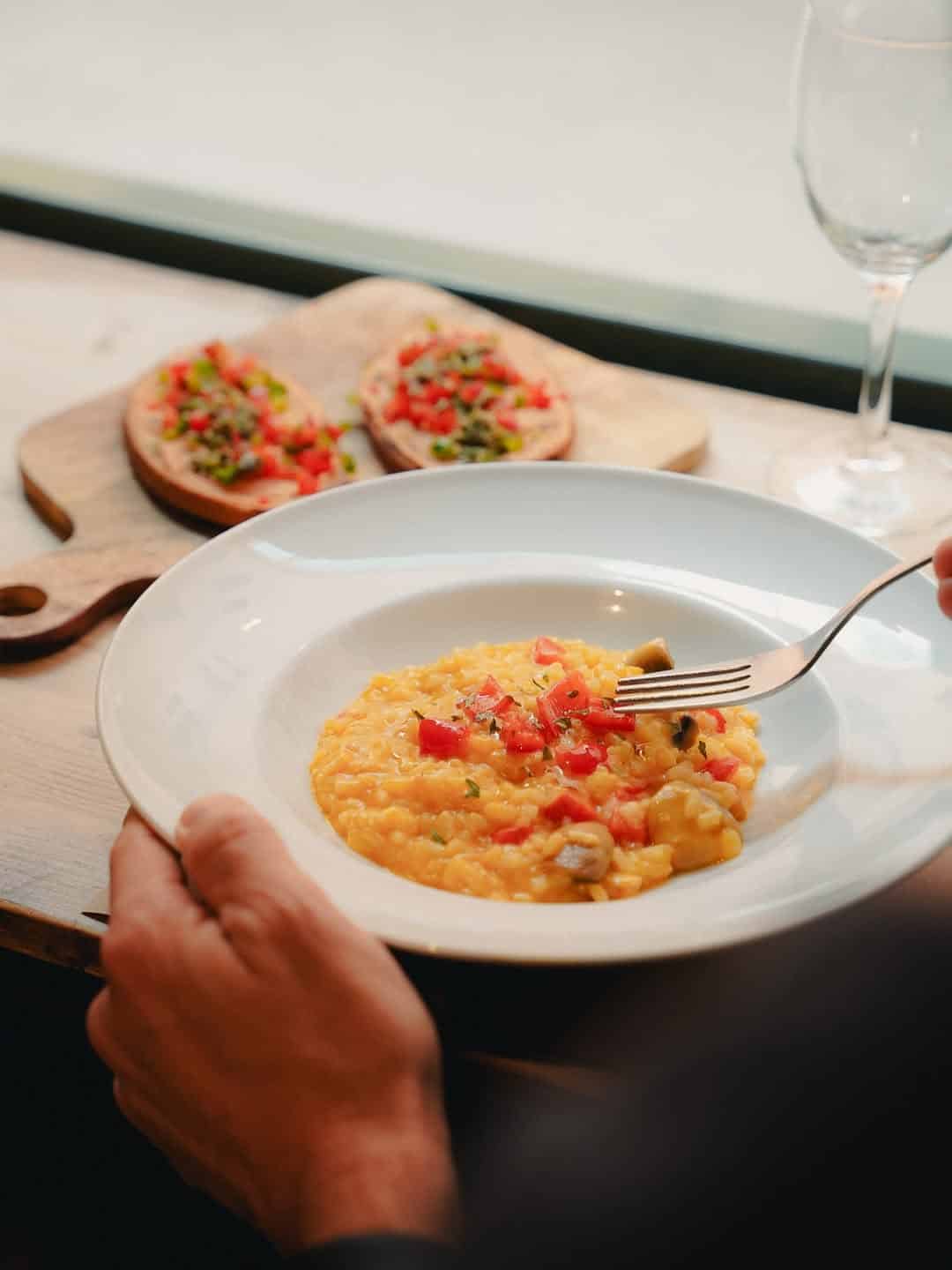 Person holding a plate of risotto with diced vegetables, perfect for a Gluten Free Barcelona experience, with bruschetta and an empty wine glass in the background.