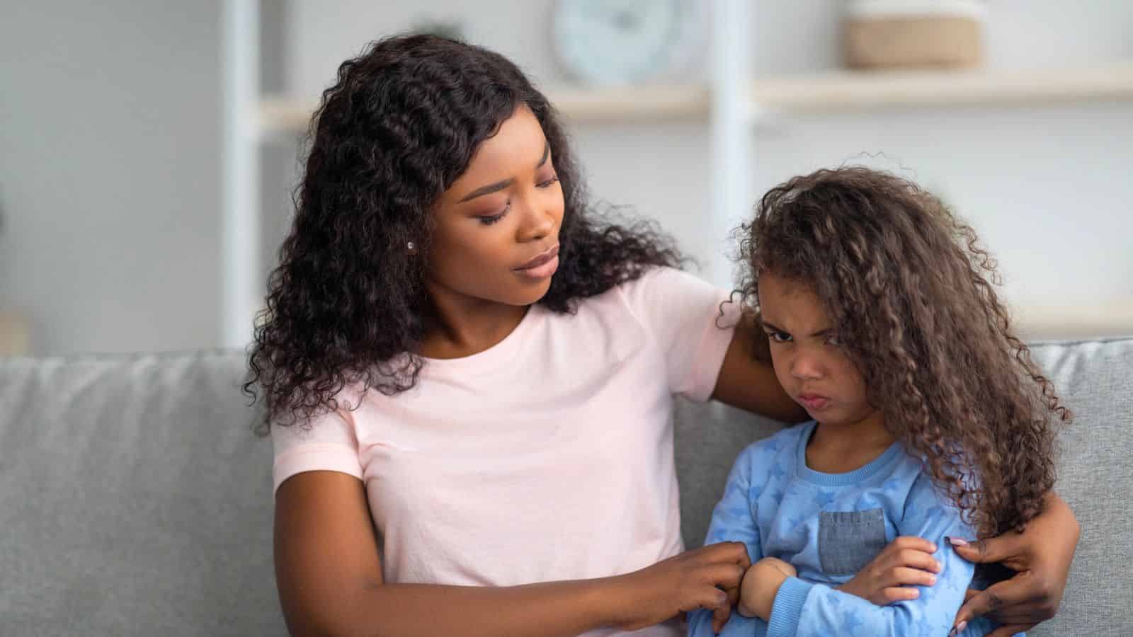 A woman comforts a young girl who looks upset, both sitting on a couch at home.