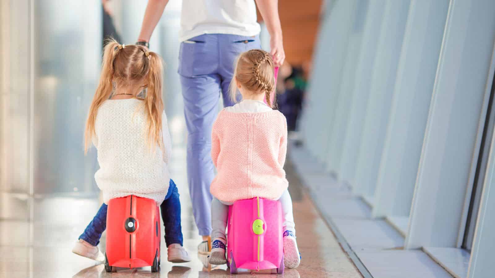 Two young girls sit on colorful suitcases as an adult walks with them in an airport corridor.