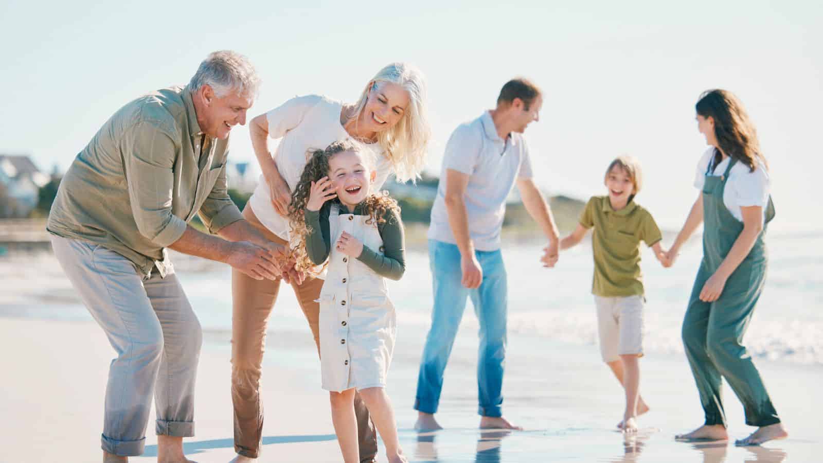 Three generations of a family laugh and play together on a sunny beach near the water.