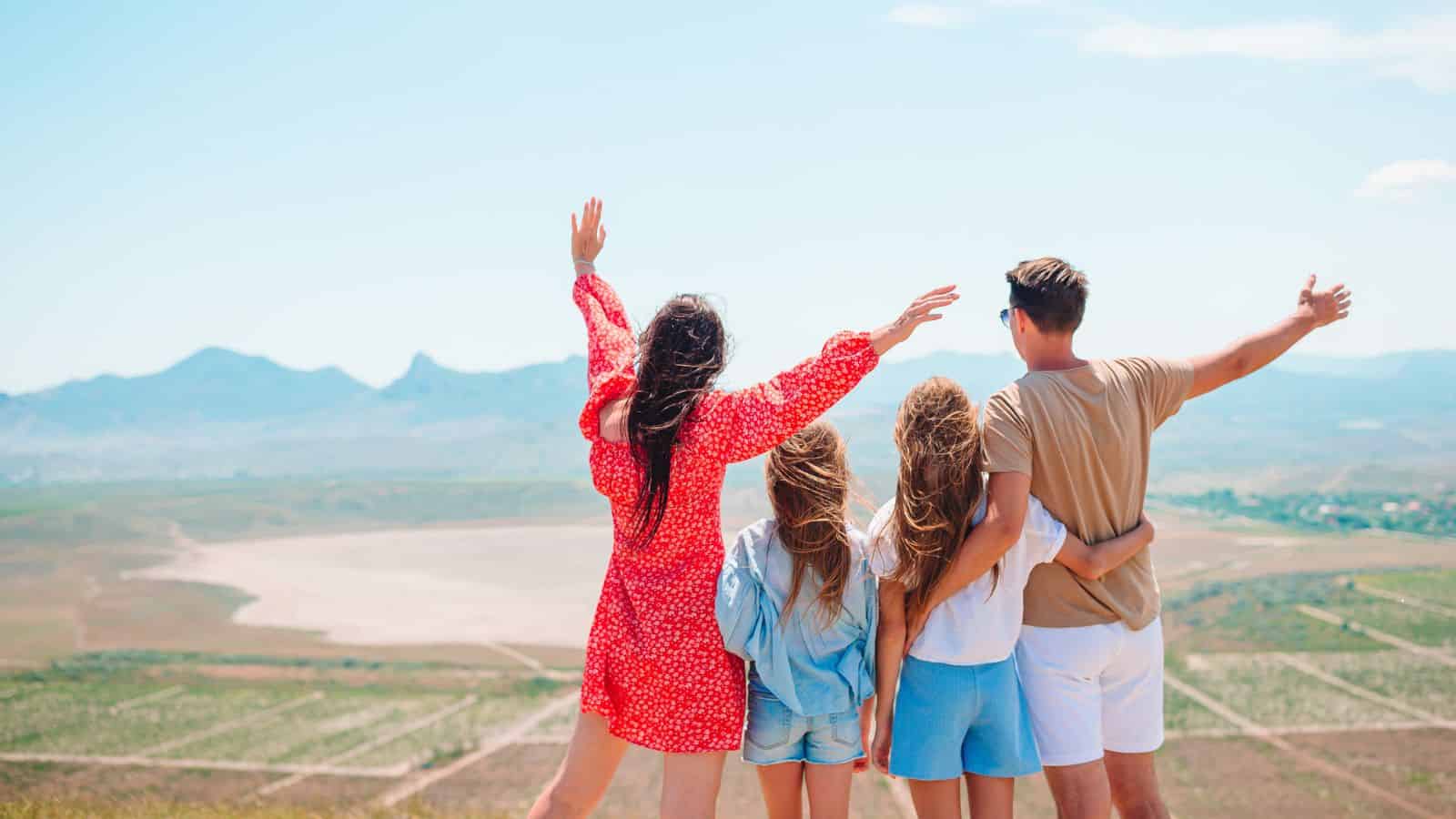 Four people stand outdoors, facing mountains, with arms raised and embracing, enjoying the scenic view.