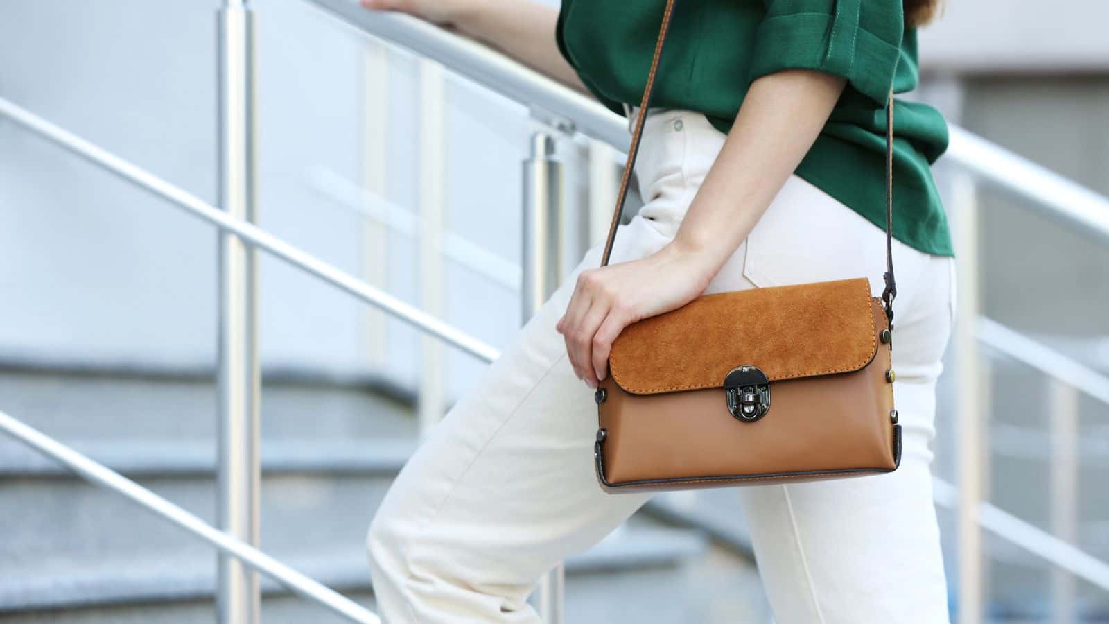 Woman in white pants and green top holding a brown crossbody bag while standing on stairs.