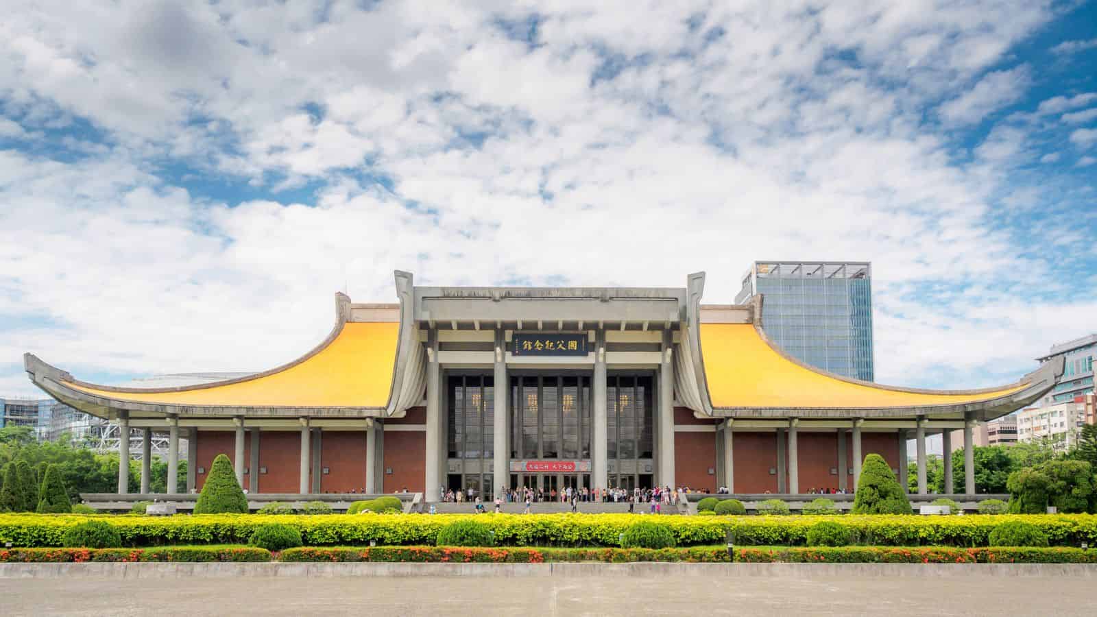 A large traditional building with a yellow curved roof and a garden in front under a blue sky with clouds.