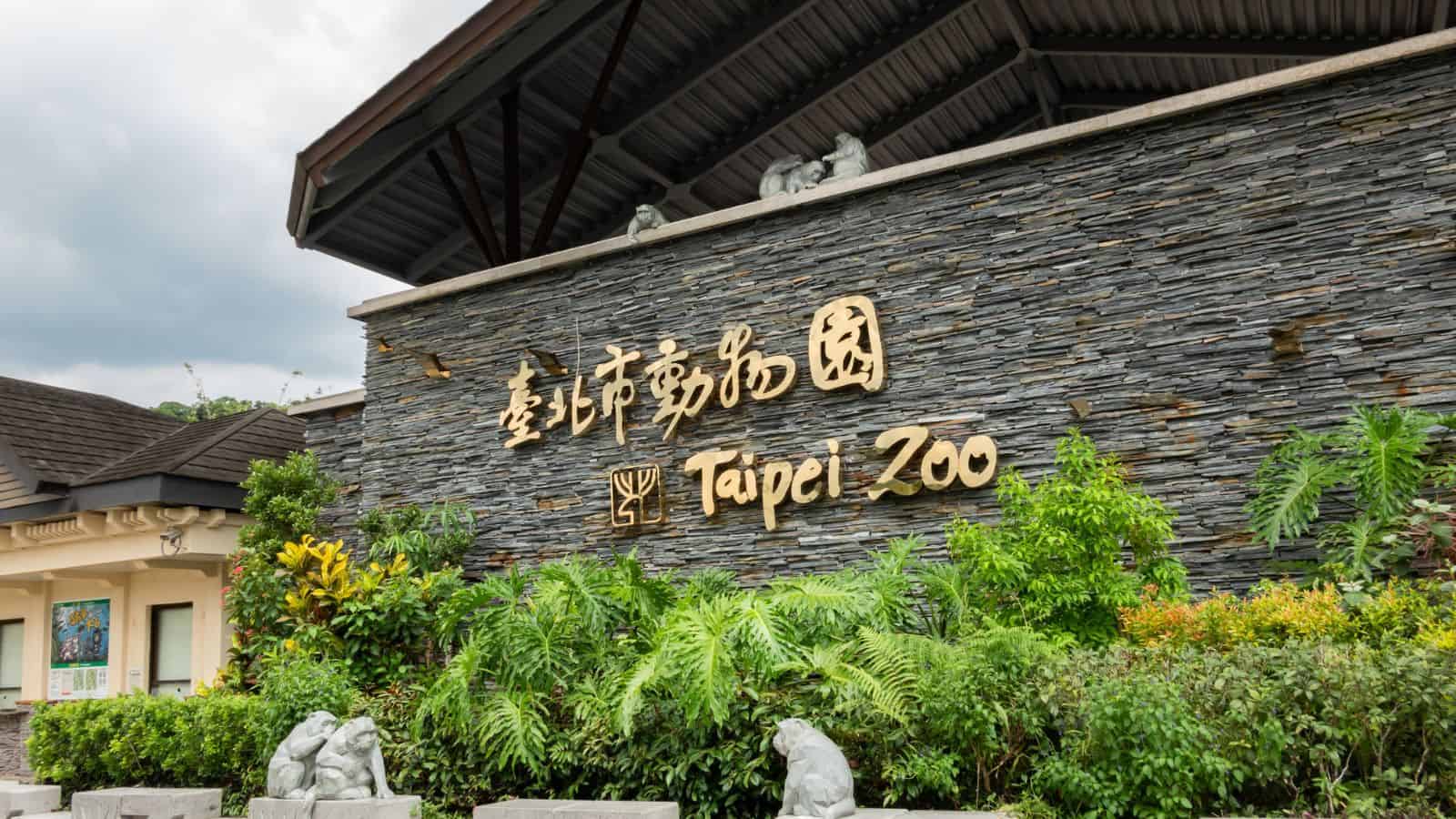 Stone wall entrance sign with "Taipei Zoo" in Chinese and English, surrounded by greenery and plants.