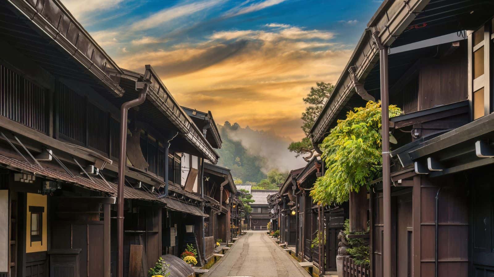 Traditional wooden Japanese buildings line a quiet street at sunset with mountains in the background.