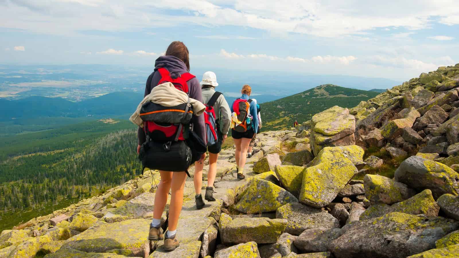 Three hikers with backpacks walk along a rocky mountain trail with scenic views and greenery in the distance.