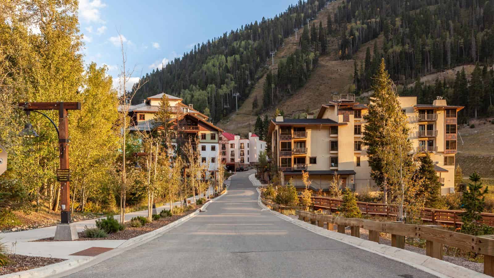 Mountain resort buildings line a quiet road, surrounded by trees and a hillside with a ski lift in the background.