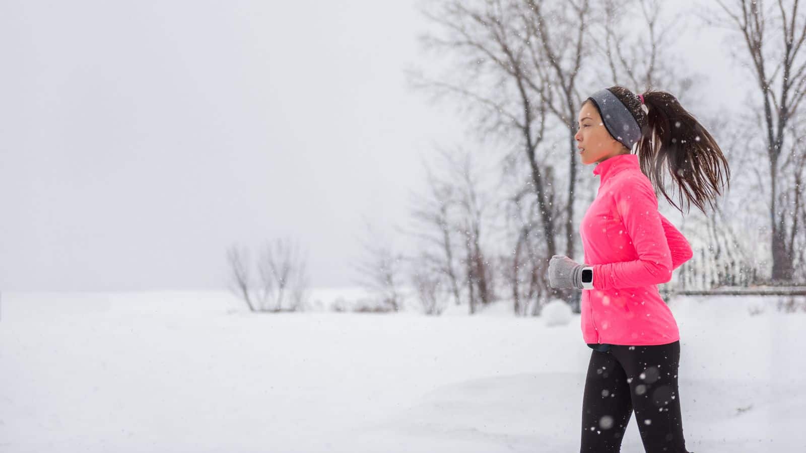 Woman jogging in the snow wearing a bright pink jacket and headband, with snowy trees in the background.