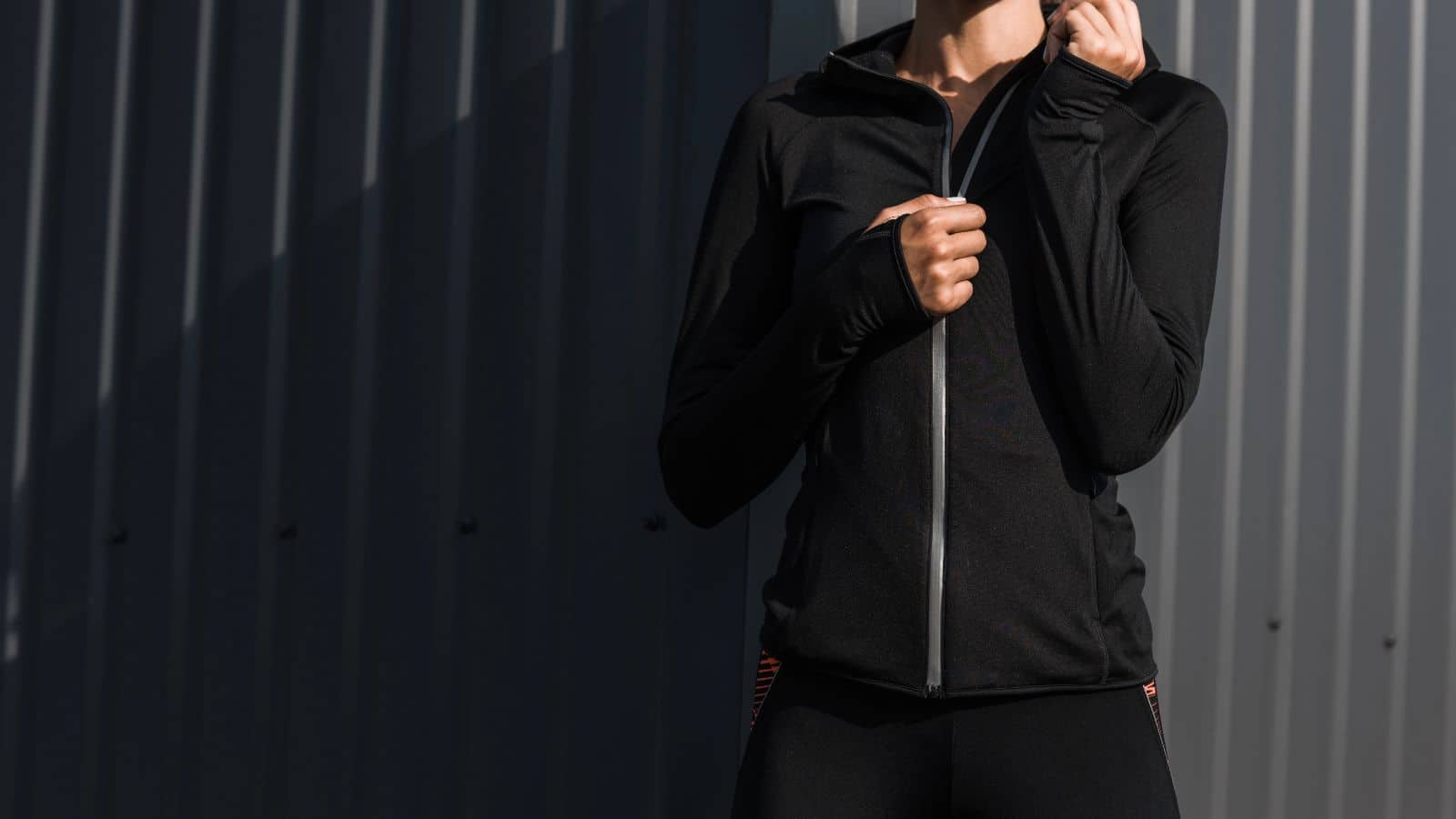 Person in a black zip-up jacket standing against a corrugated metal wall, face not visible.