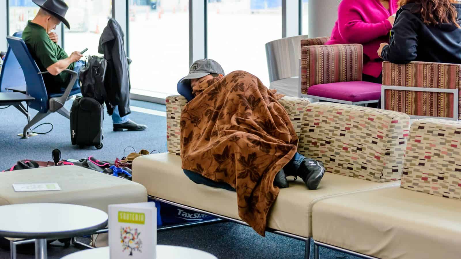 A person sleeps on an airport bench under a brown blanket while others sit nearby.