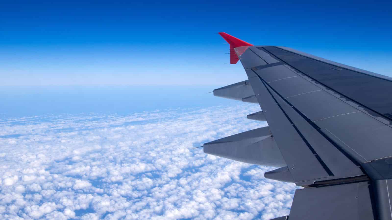 View from airplane window showing wing and clouds below against a bright blue sky.
