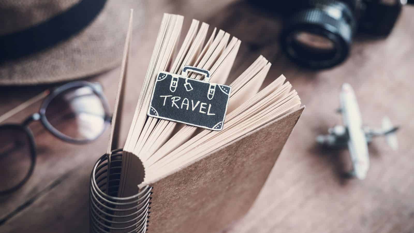 Spiral notebook with a "Travel" suitcase clip, glasses, hat, camera, and toy airplane on a wooden table.