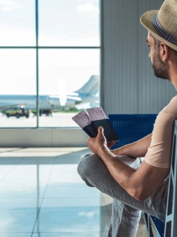 Man in hat sitting at airport gate, holding tickets and passport, looking at plane outside window.