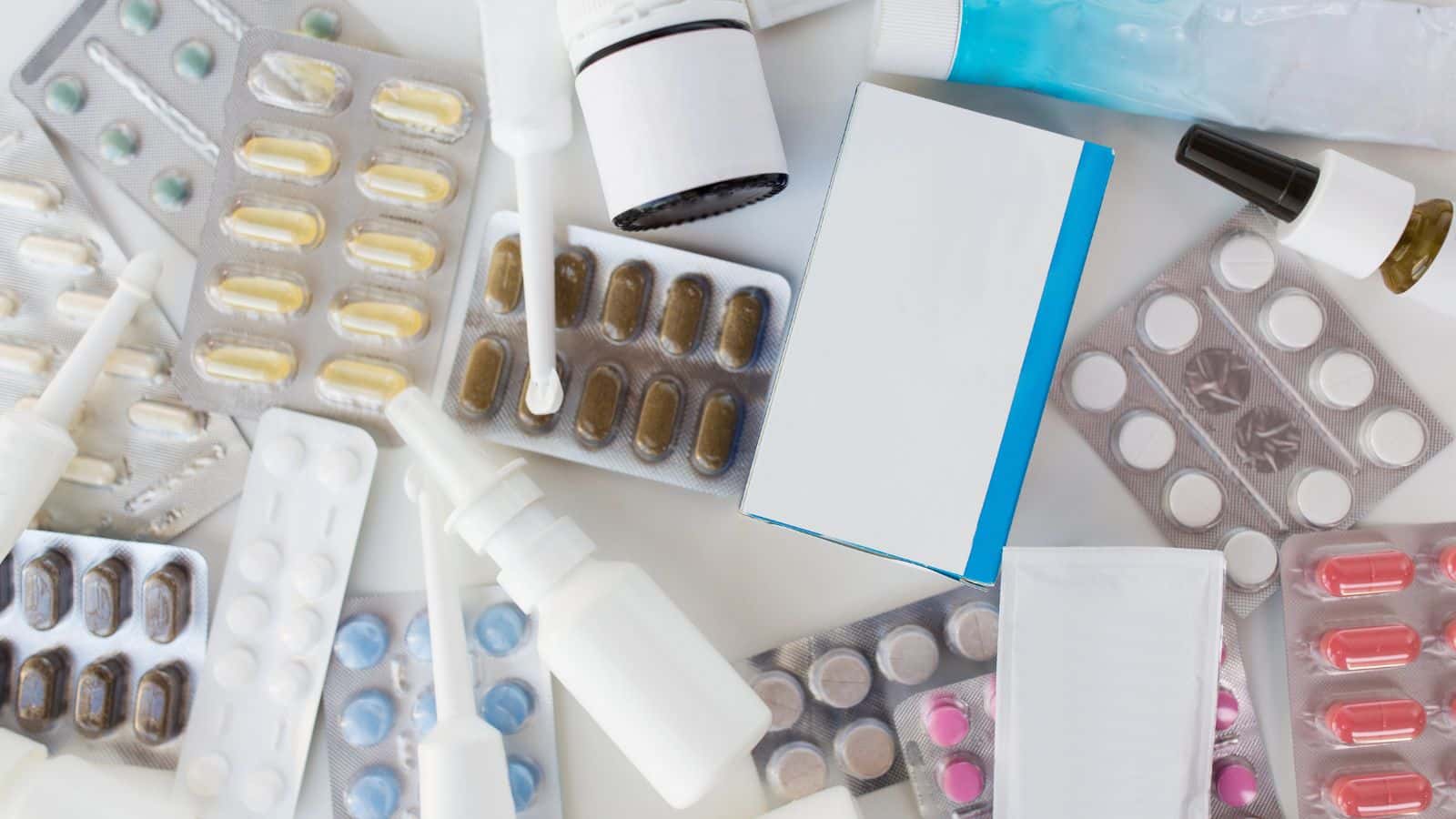 Various blister packs, pill bottles, nasal sprays, and medicine boxes scattered on a white surface.