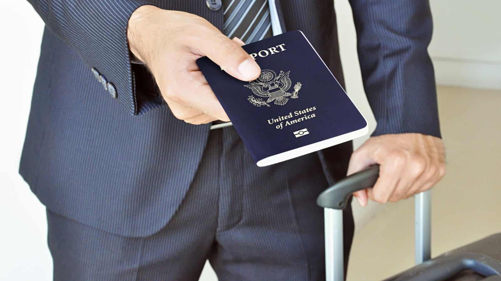 Man in suit holding a U.S. passport and rolling a suitcase, ready for travel.