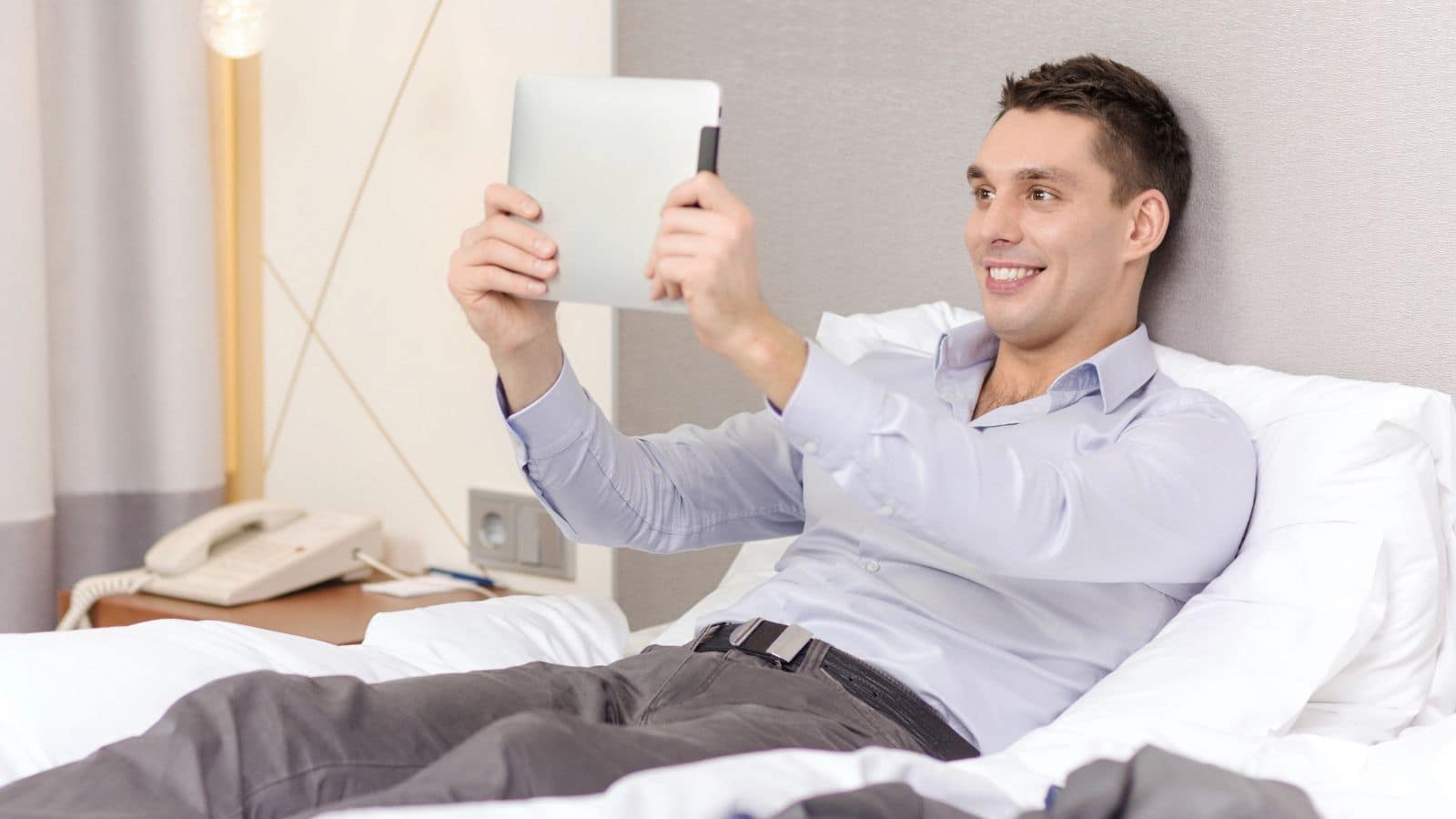 Smiling man lying on a bed, holding and looking at a tablet device.