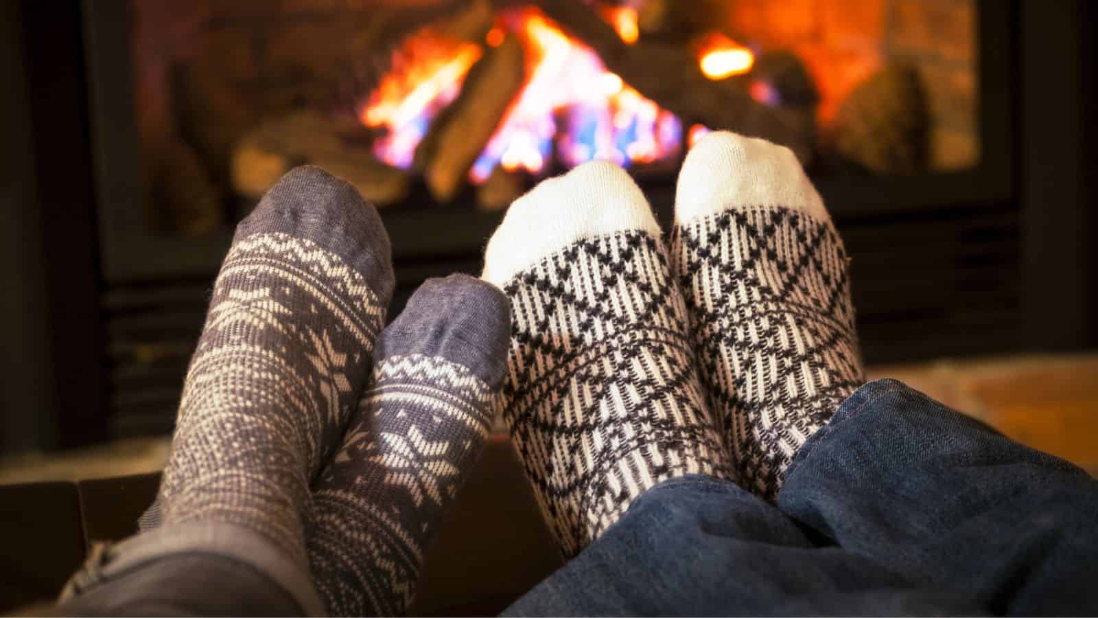 Two people wearing patterned socks relax by a cozy fireplace, their feet close and legs stretched out.