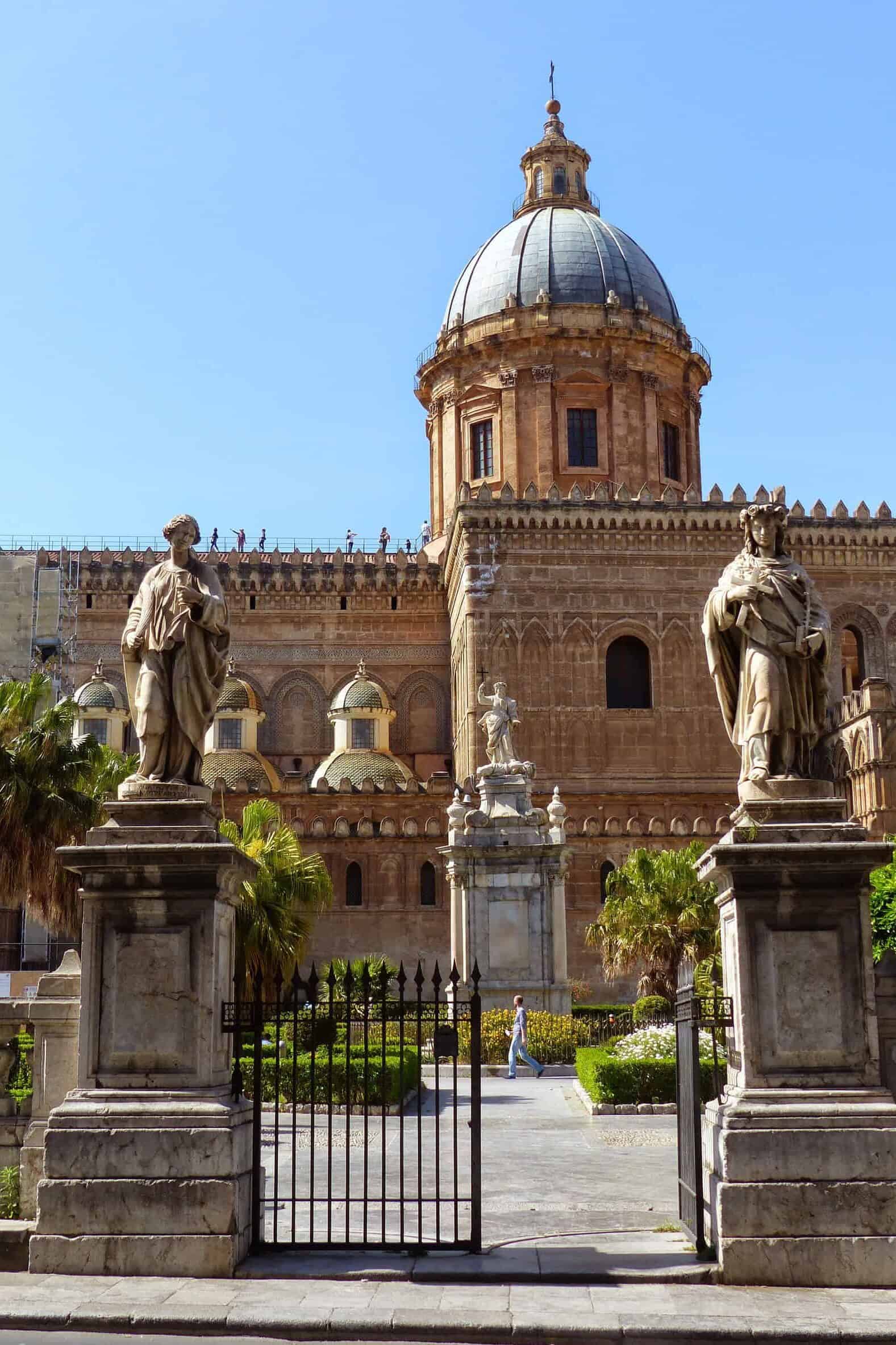 Historic cathedral with a large dome, flanked by two statues and a black iron gate in the foreground&mdash;a must-see if you're wondering what to do in Palermo for a day.