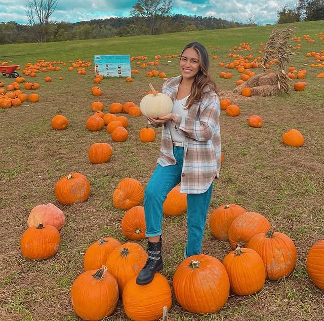 A woman in a plaid shirt holds a white pumpkin, standing among orange pumpkins in one of the best pumpkin patches.
