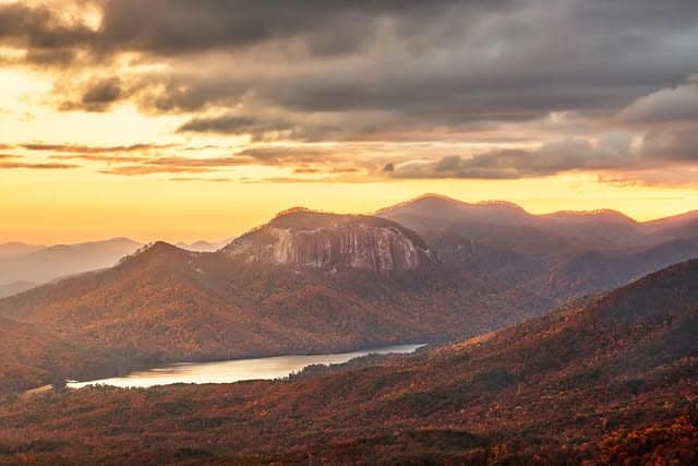 Sunset over rolling mountains with autumn foliage, a large rock face, and a lake nestled in the valley below—one of the best mountain getaways for nature lovers.