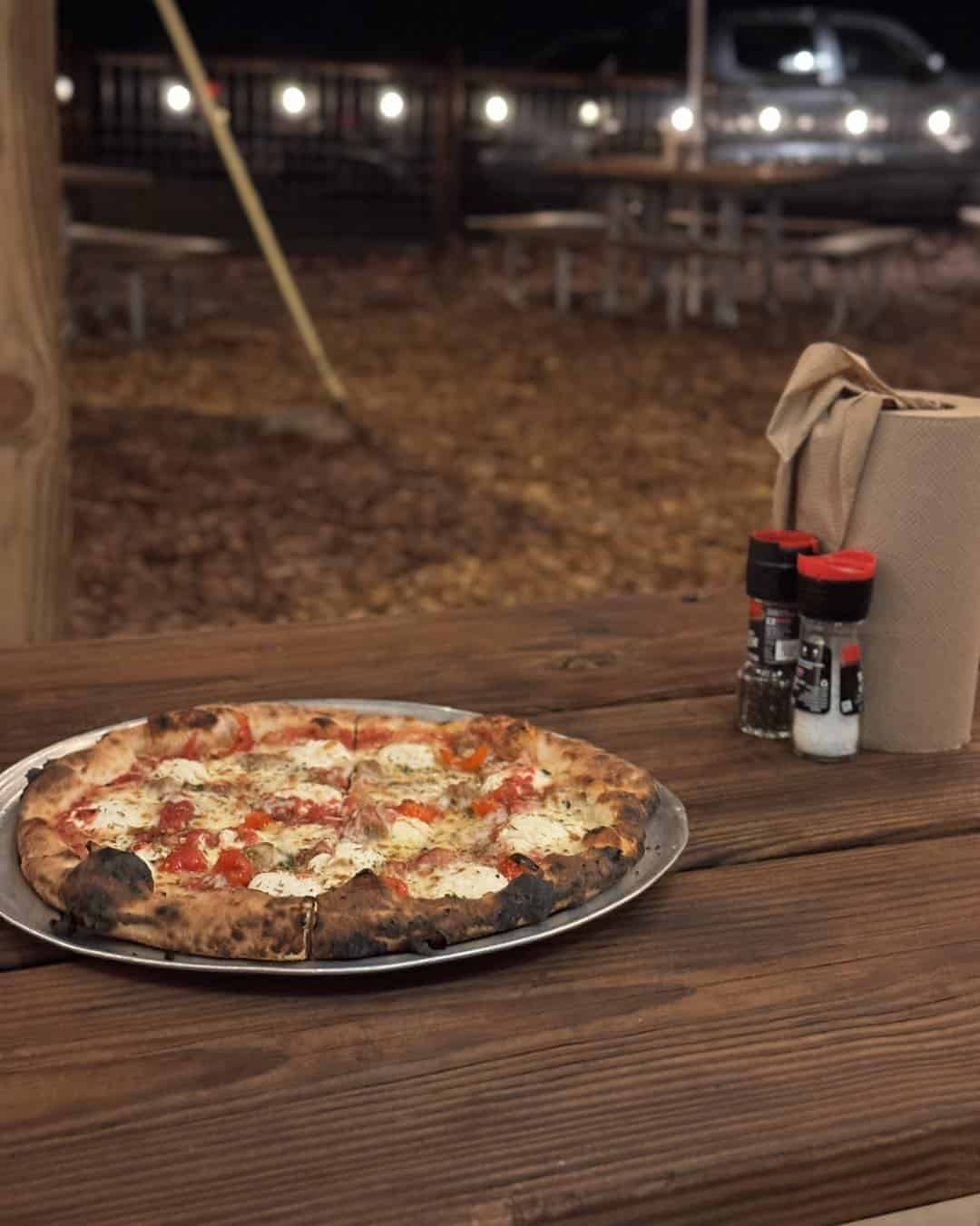 A pizza on a wooden table outdoors, with napkins and seasonings, picnic tables in the background—a perfect meal to savor during one of the best mountain getaways.