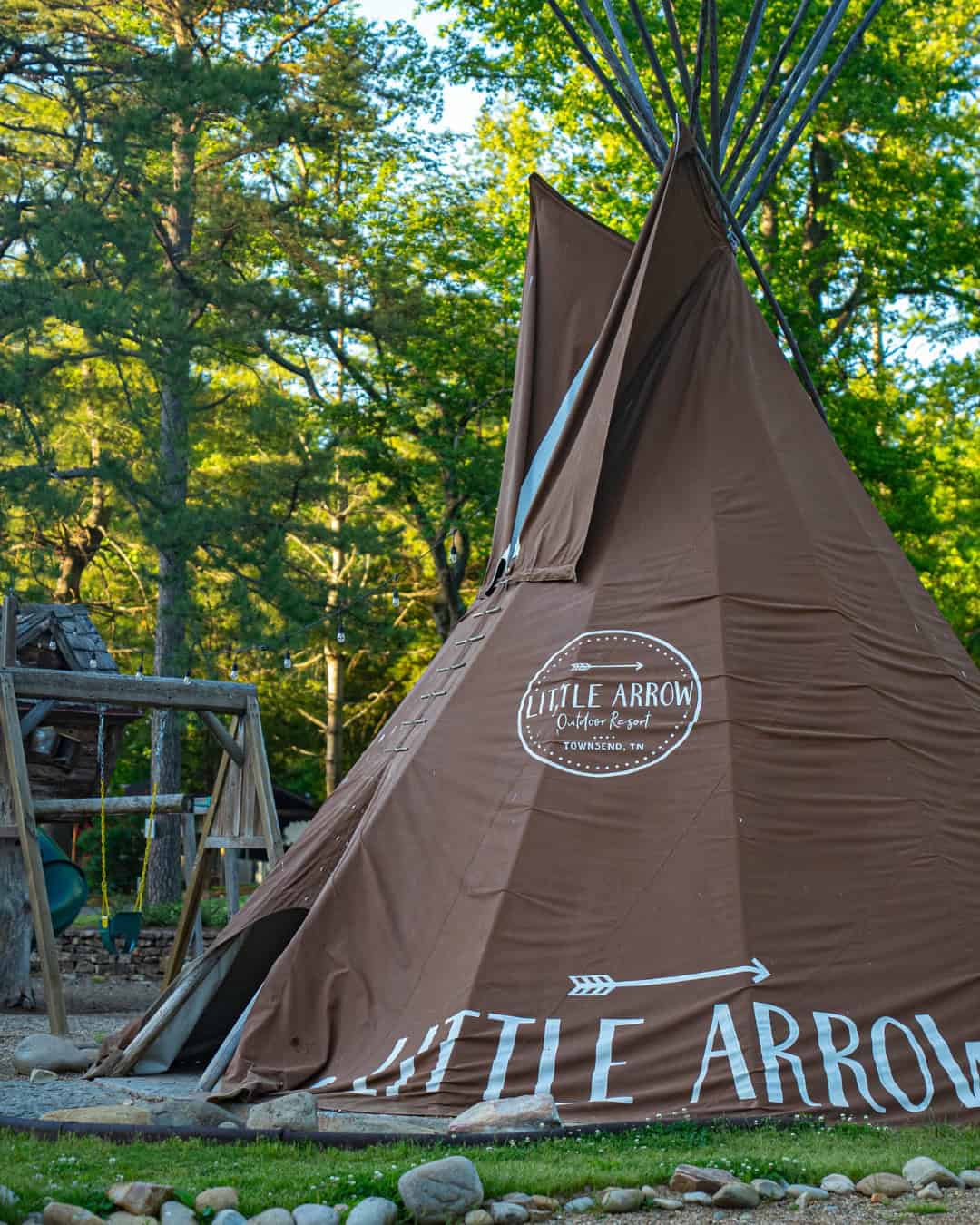 A large brown teepee labeled "Little Arrow" stands among trees at an outdoor campsite, offering one of the best mountain getaways for nature lovers.
