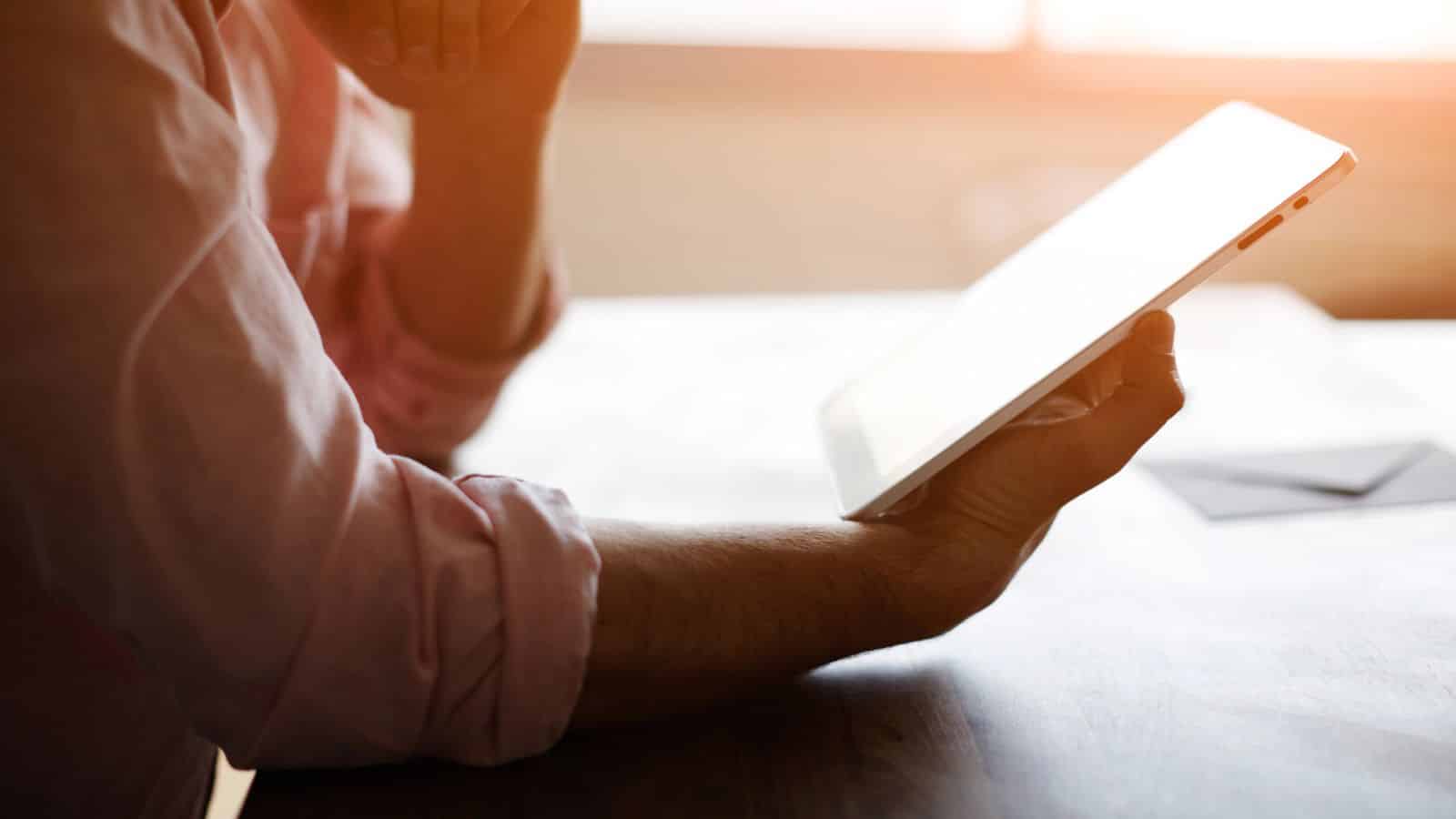 Person in a pink shirt holding and looking at a tablet at a sunlit desk.