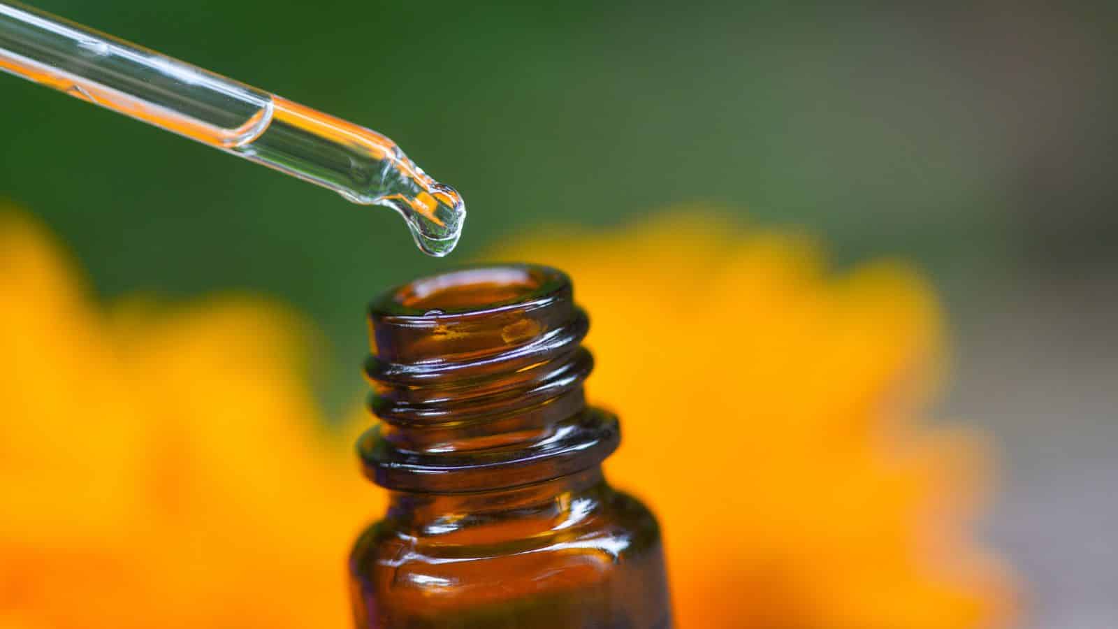 A dropper with liquid above a small amber bottle, with an orange flower blurred in the background.