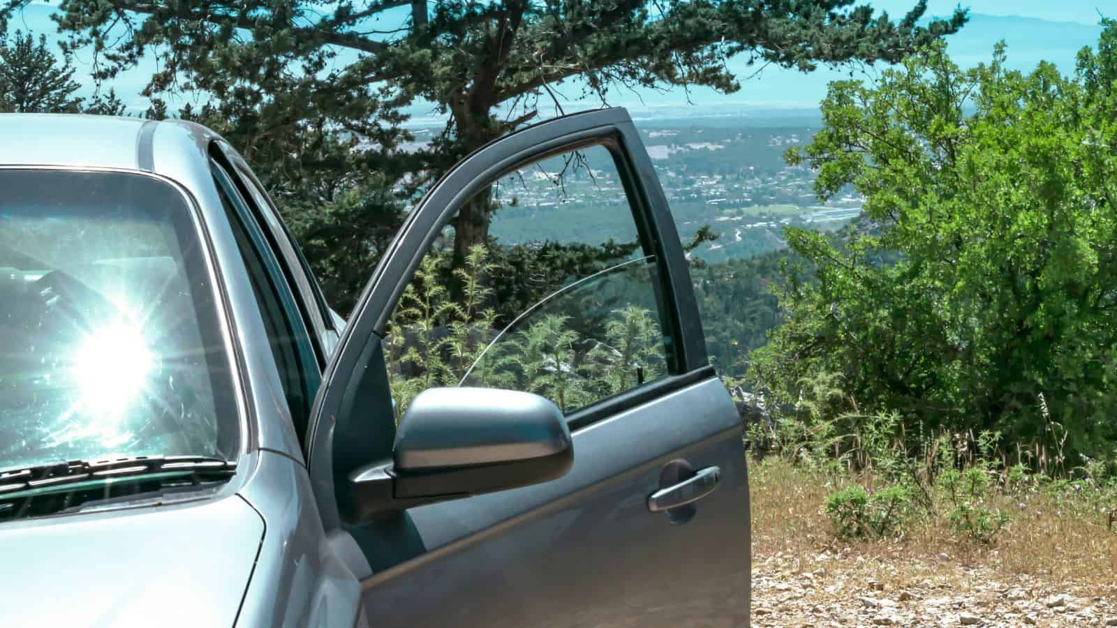 A silver car with an open door is parked by a scenic overlook with trees and a view in the background.