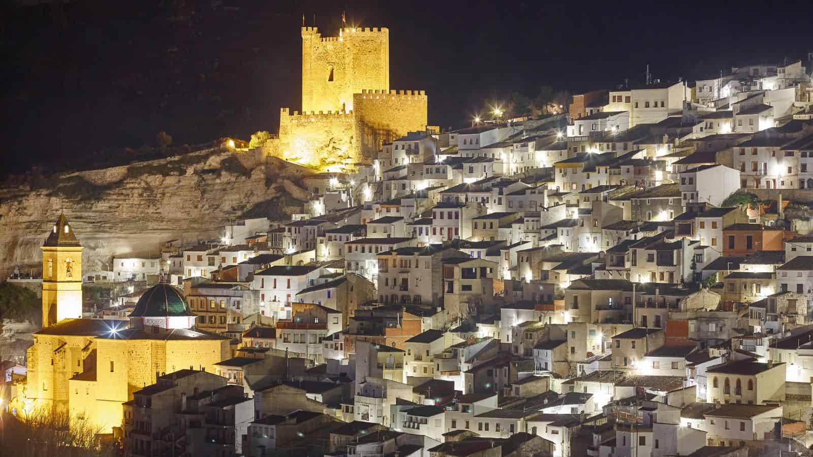 A hillside town at night with illuminated white houses and a lit castle on top.
