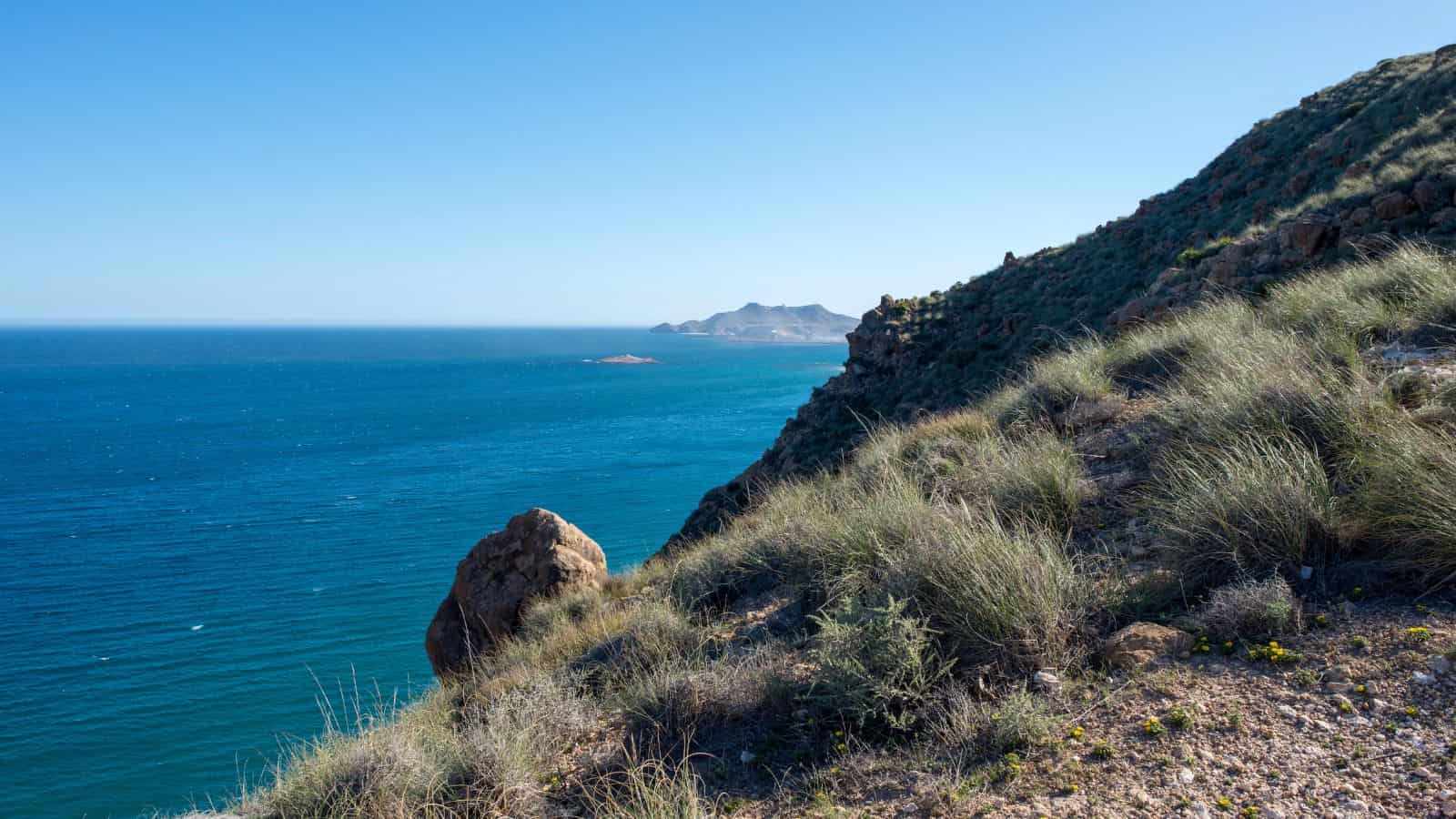 Rocky hillside with grass overlooking a blue sea, distant mountains visible under a clear sky.