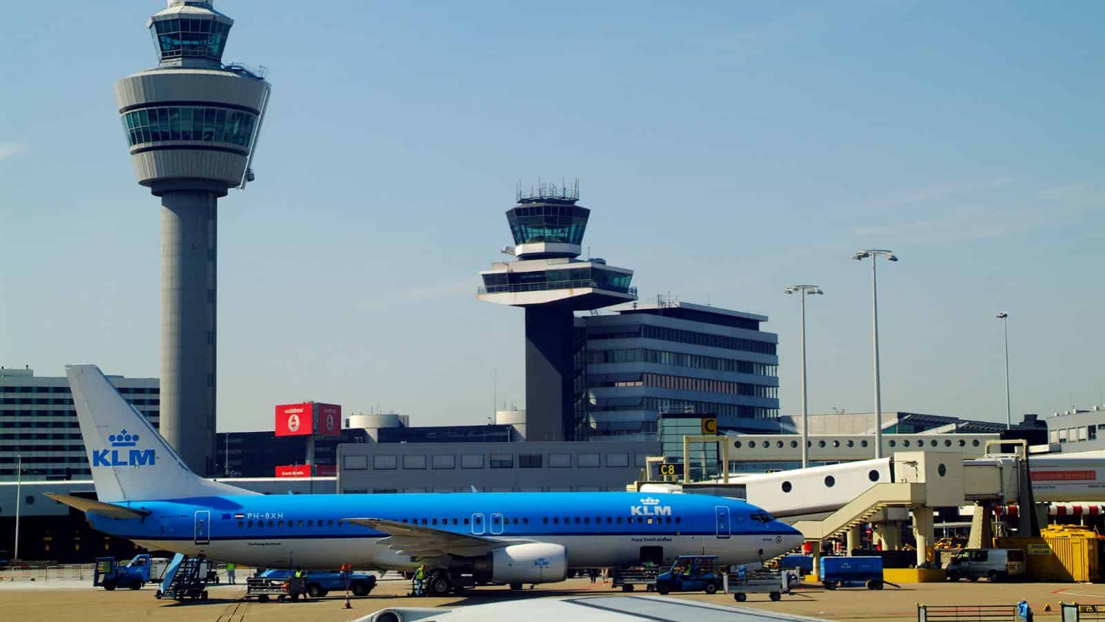 A KLM airplane parked at an airport gate with control towers and terminal buildings in the background.