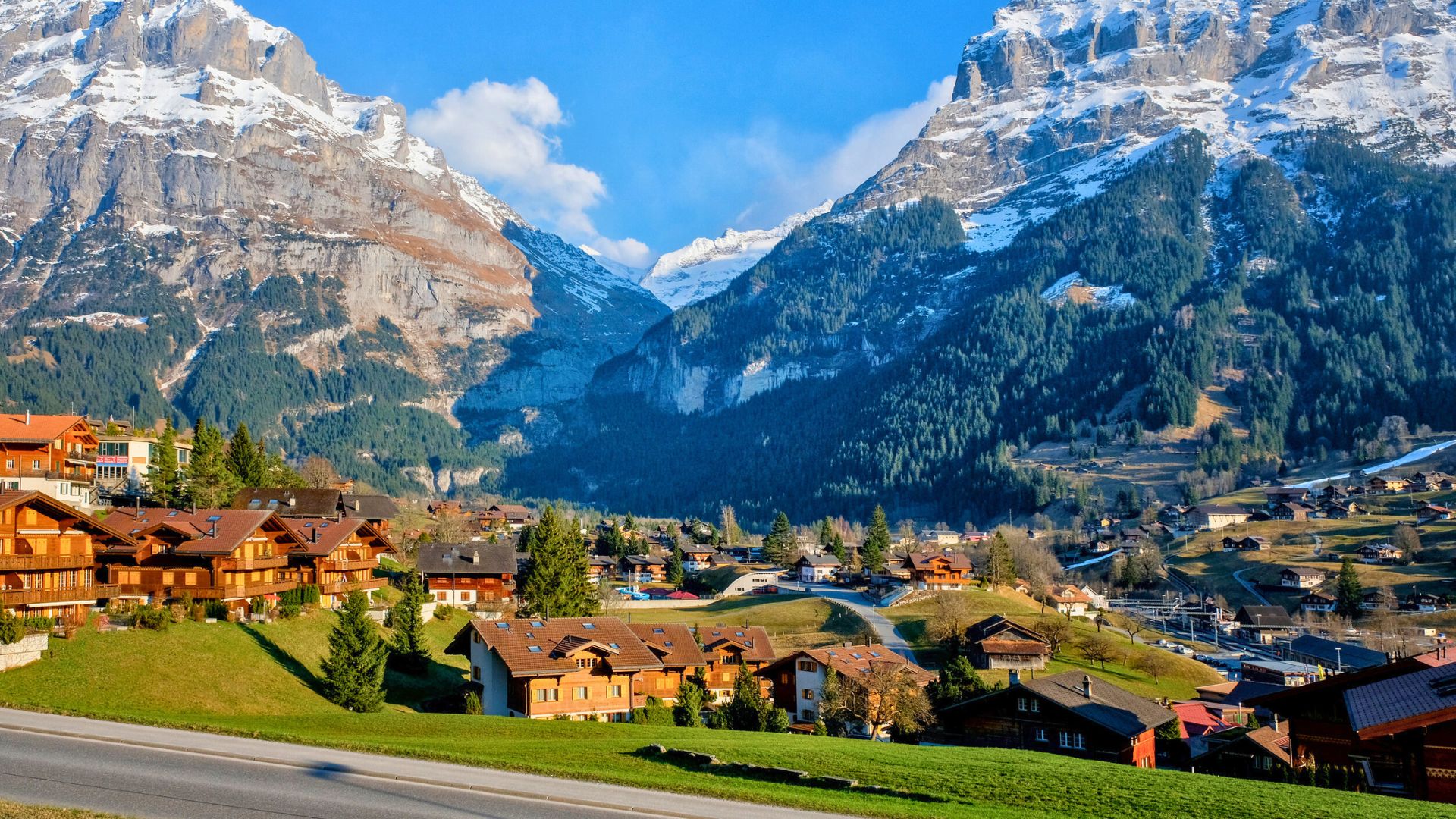 Chalets in a green valley surrounded by snow-capped mountains under a clear blue sky.