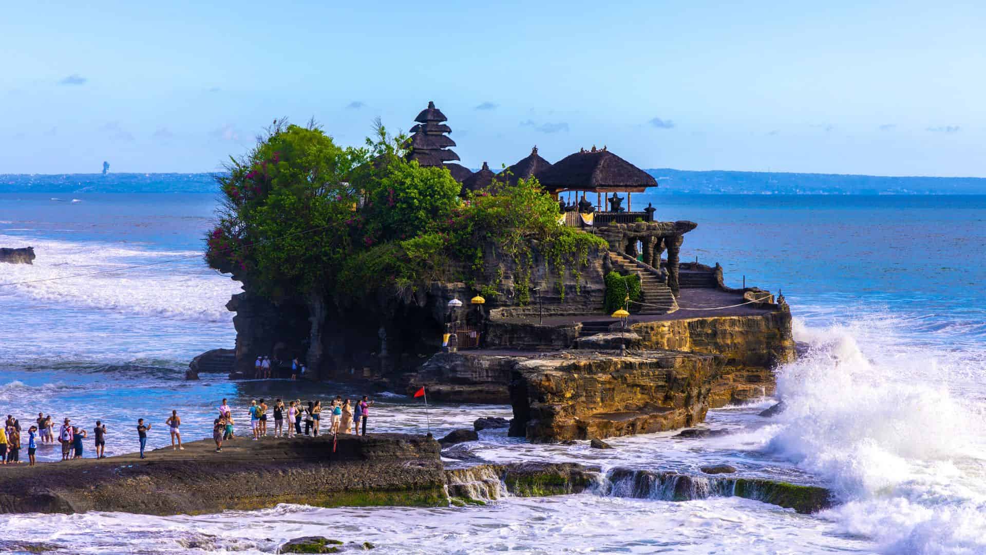 Tanah Lot temple sits on a rocky island as waves crash, with tourists walking along the shore in Bali, Indonesia.