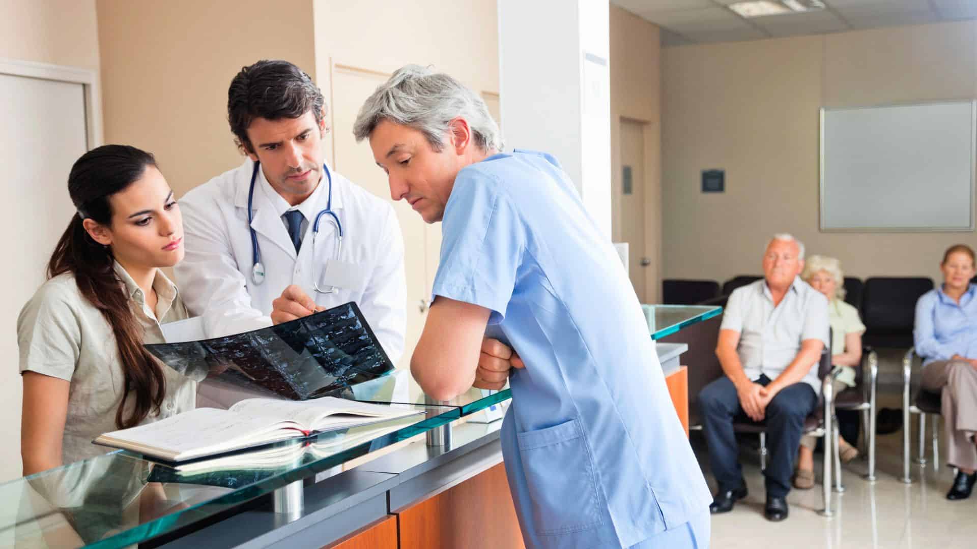 Three medical professionals review an X-ray at a reception desk; patients wait in chairs in the background.