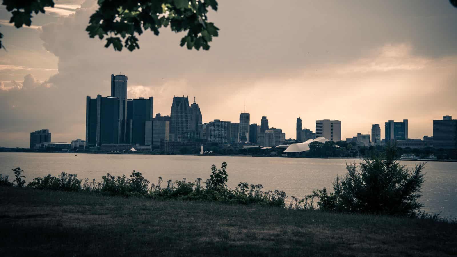 A city skyline at dusk, viewed across a river with trees and grass in the foreground.