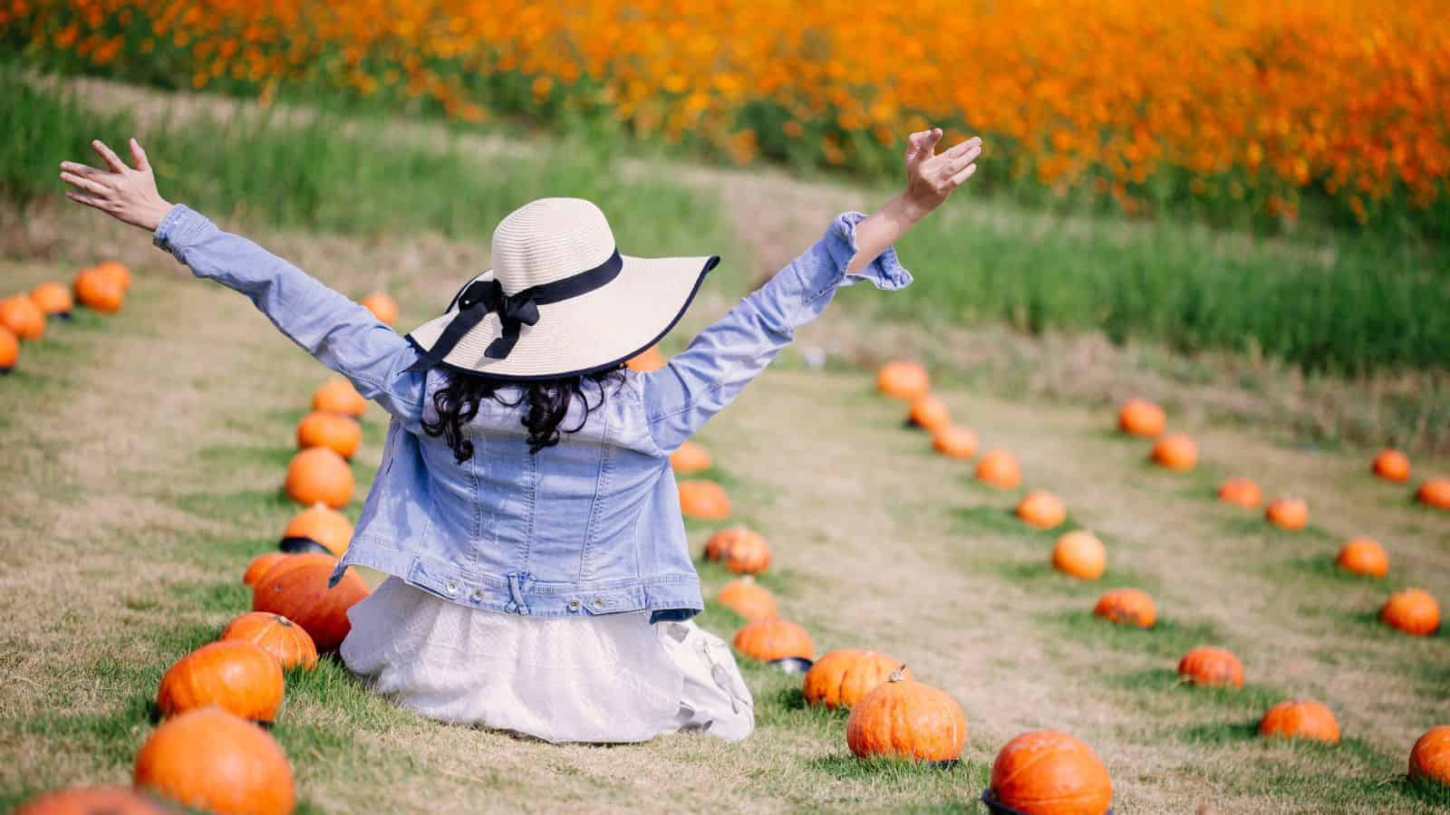 Woman in a sunhat sitting among rows of pumpkins, arms raised, facing a field of orange flowers.
